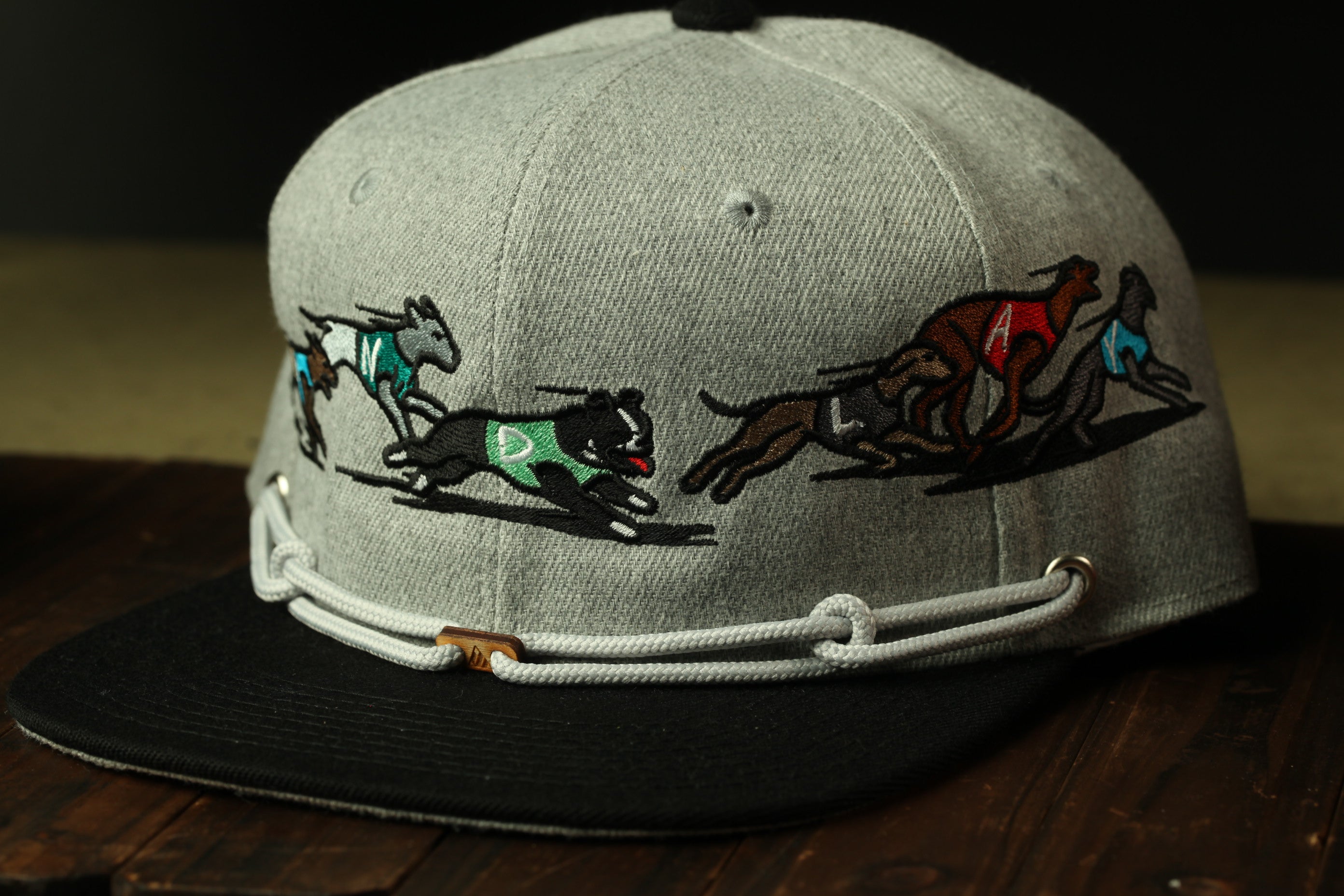 A gray baseball cap with a black brim and a white band, featuring a graphic design of three dogs running in a race.