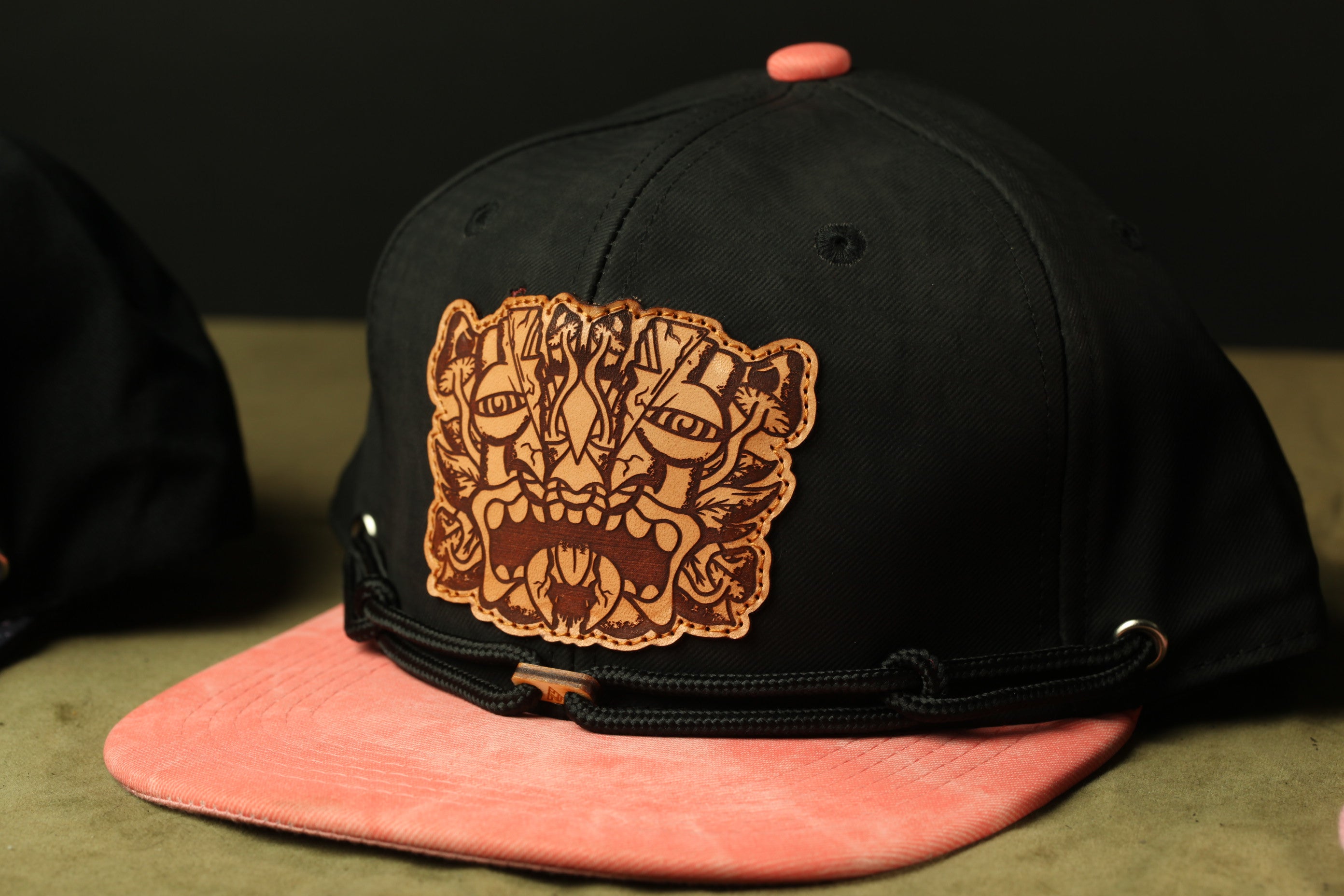 A black baseball cap with a pink brim and a detailed leather patch featuring a stylized face design.