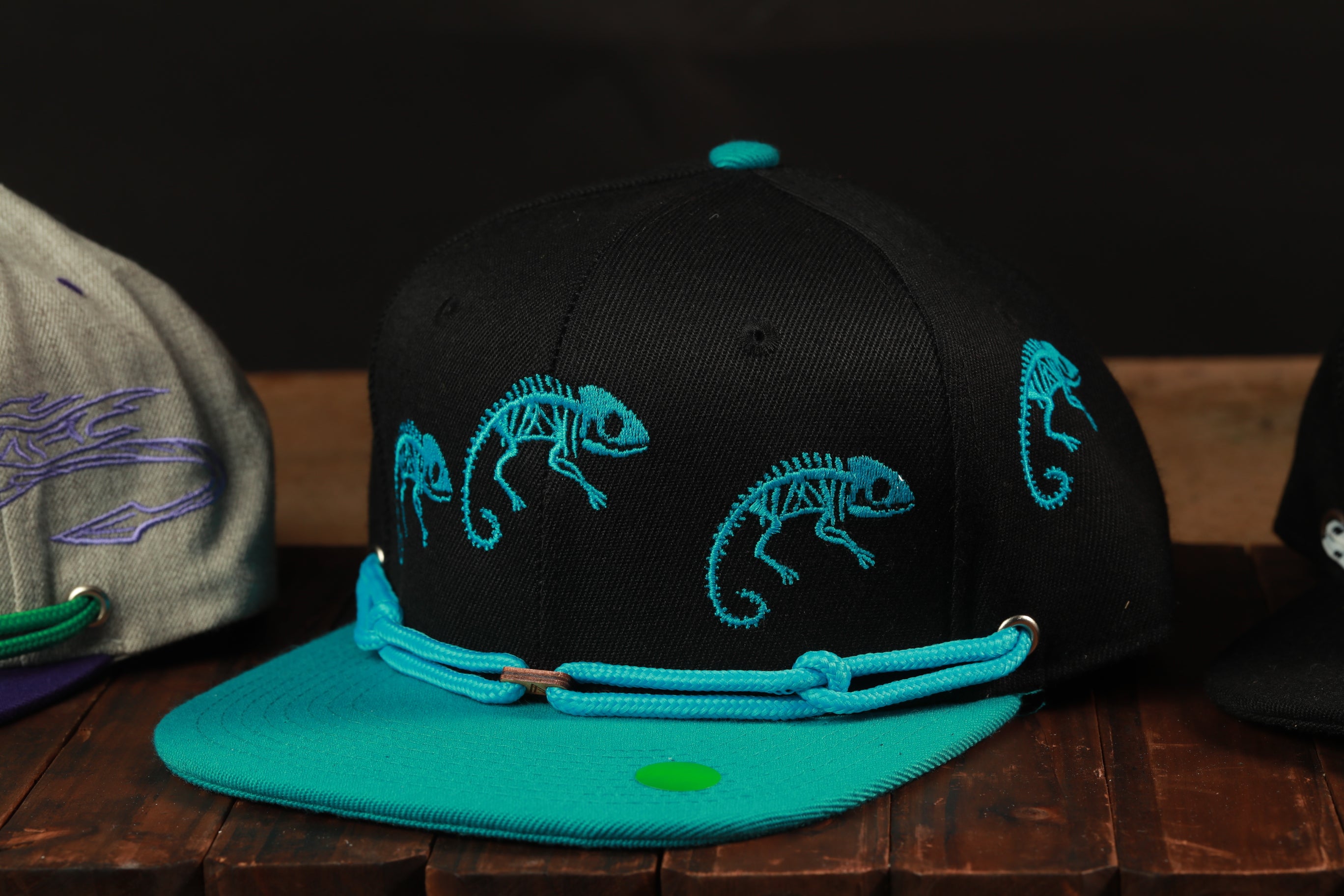 A close-up image of a black baseball cap with blue embroidered chameleons on the front, displayed on a wooden surface.