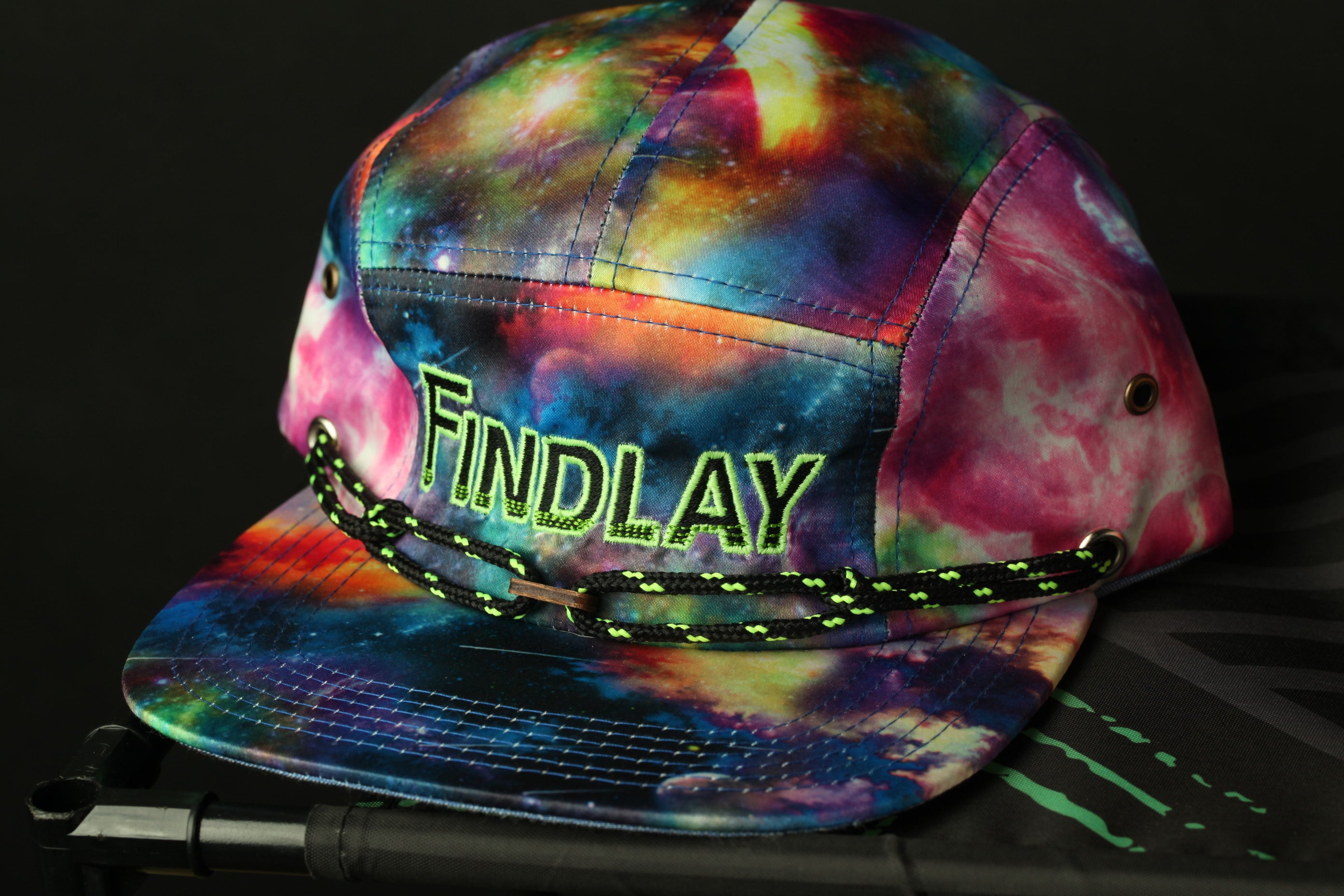 A colorful, tie-dye baseball cap with the word "FINDlay" printed on it, sitting on a surface with a black and green pattern.