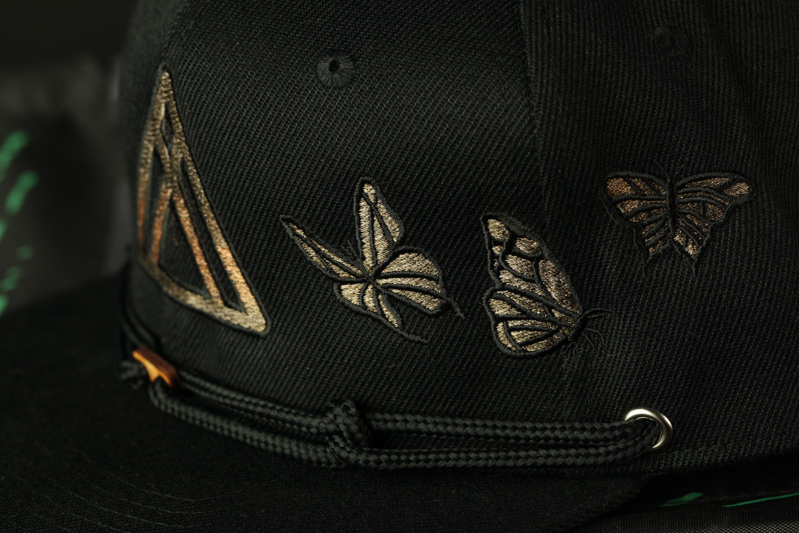 A close-up view of a black baseball cap with gold embroidered butterflies and a triangular logo.