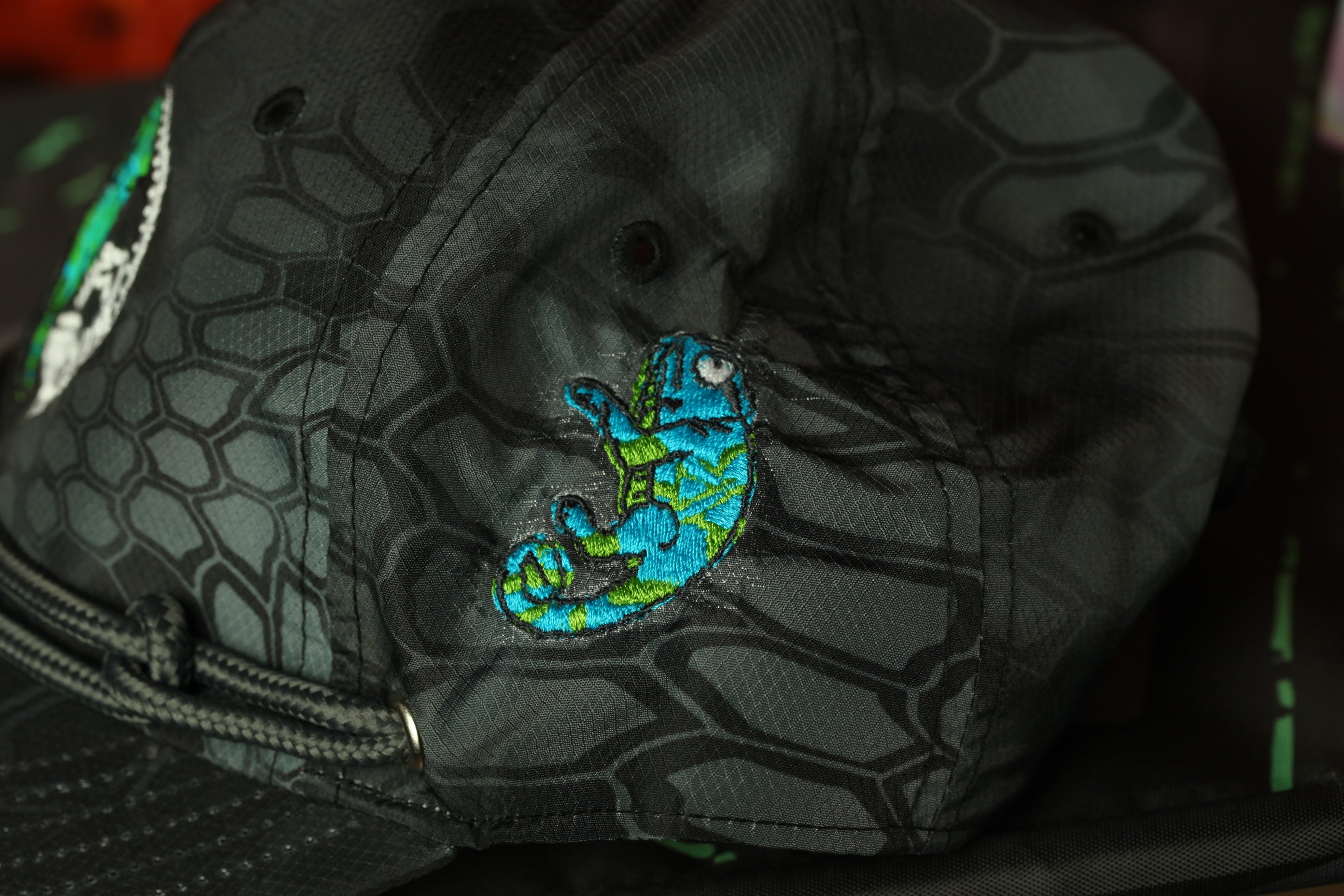 A close-up view of a black baseball cap with a unique pattern and a small embroidered fish design on the front.