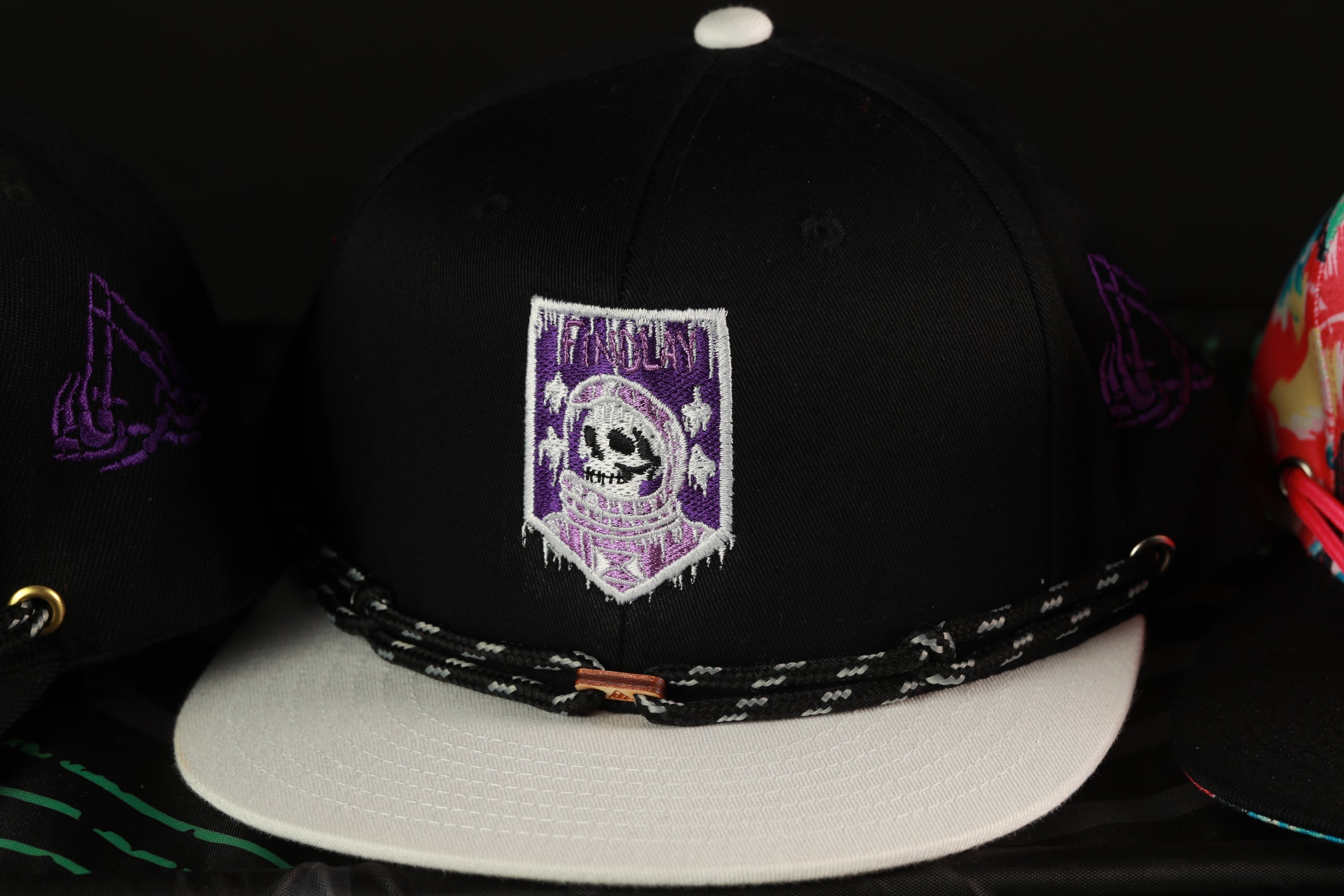 A close-up view of a black baseball cap with a purple and white skull embroidered on it, along with a black and white braided strap.