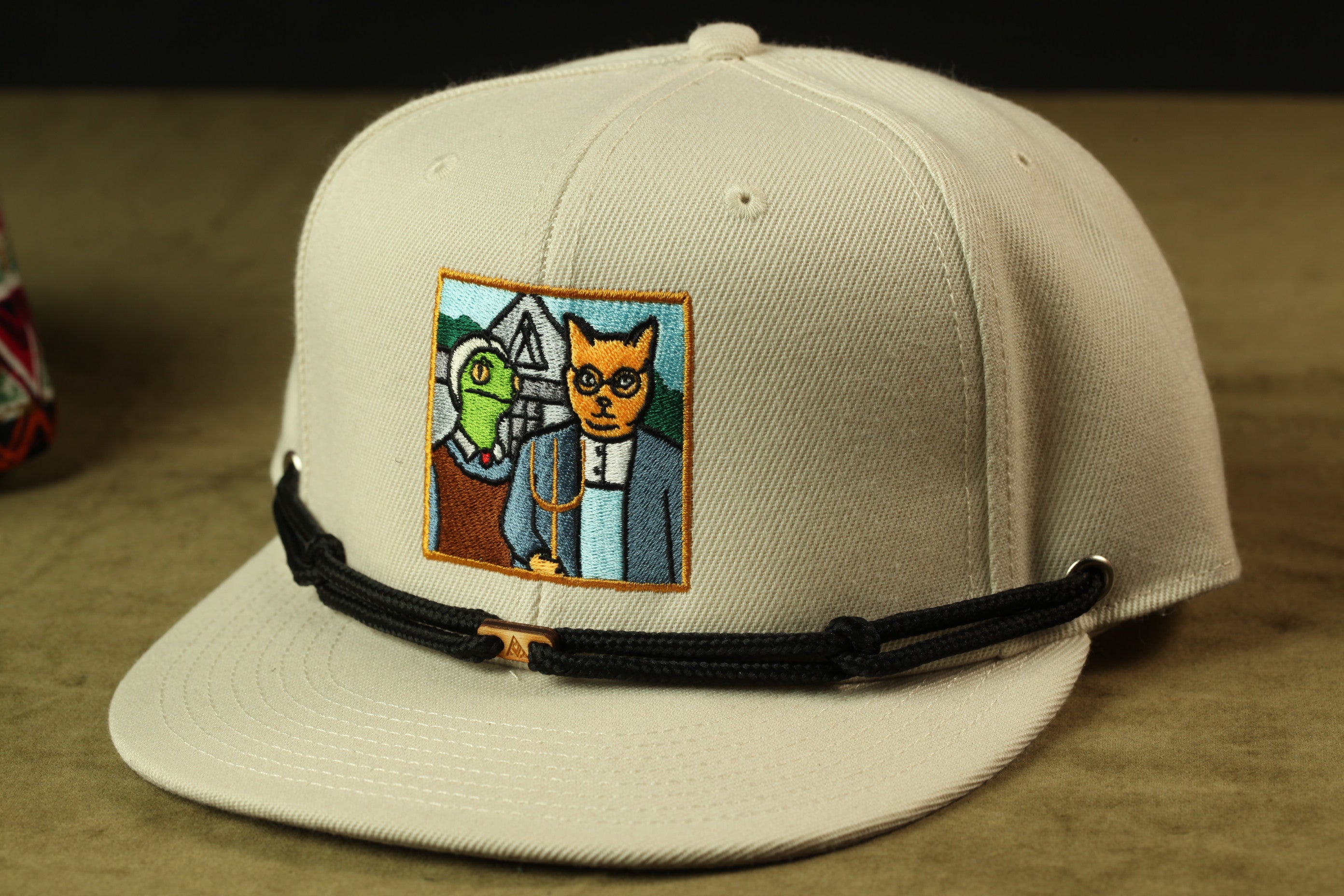 A beige baseball cap with a colorful embroidered patch depicting a cat and a bird.