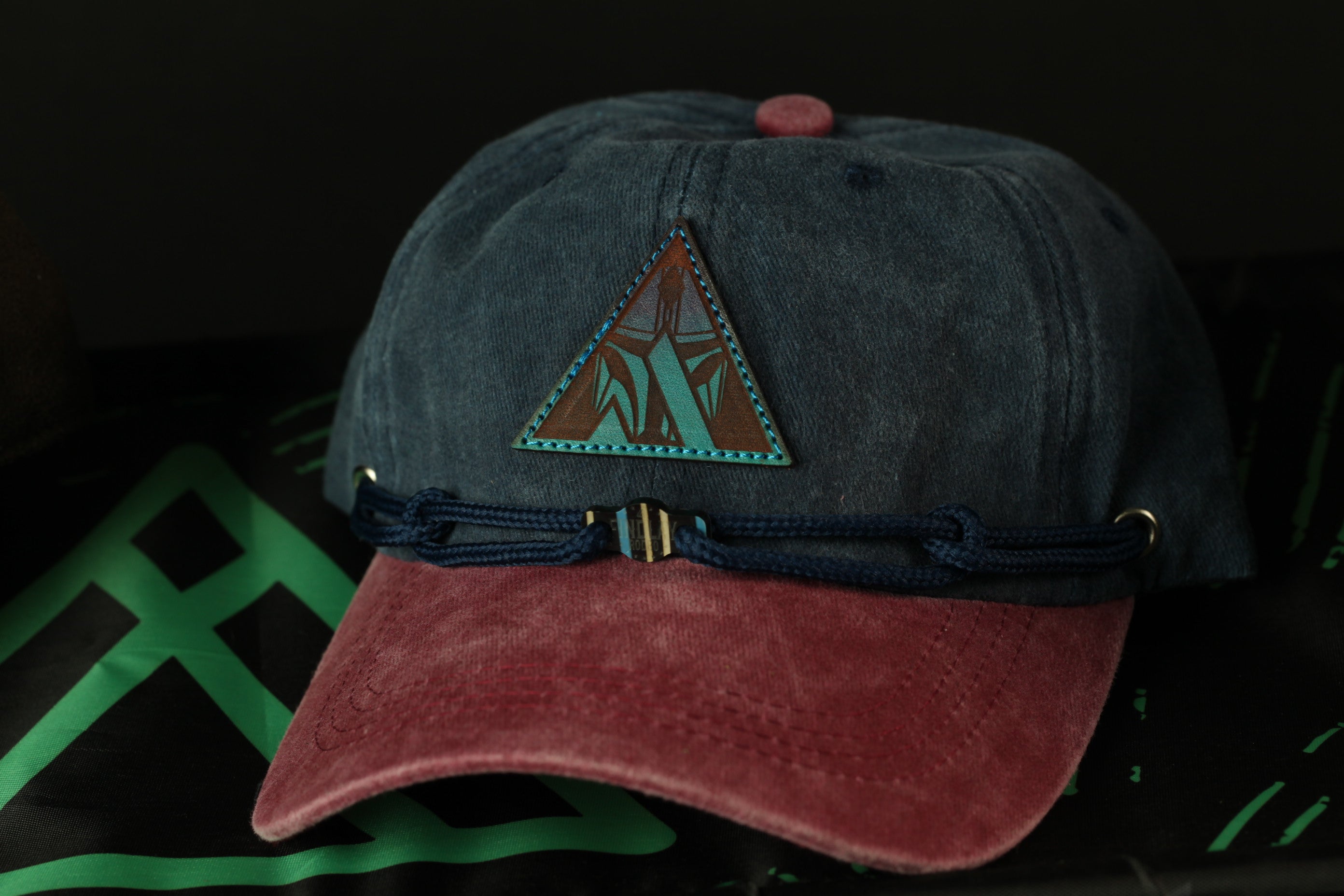 A dark blue baseball cap with a red brim and a triangular patch on the front, resting on a black surface with a green geometric pattern.