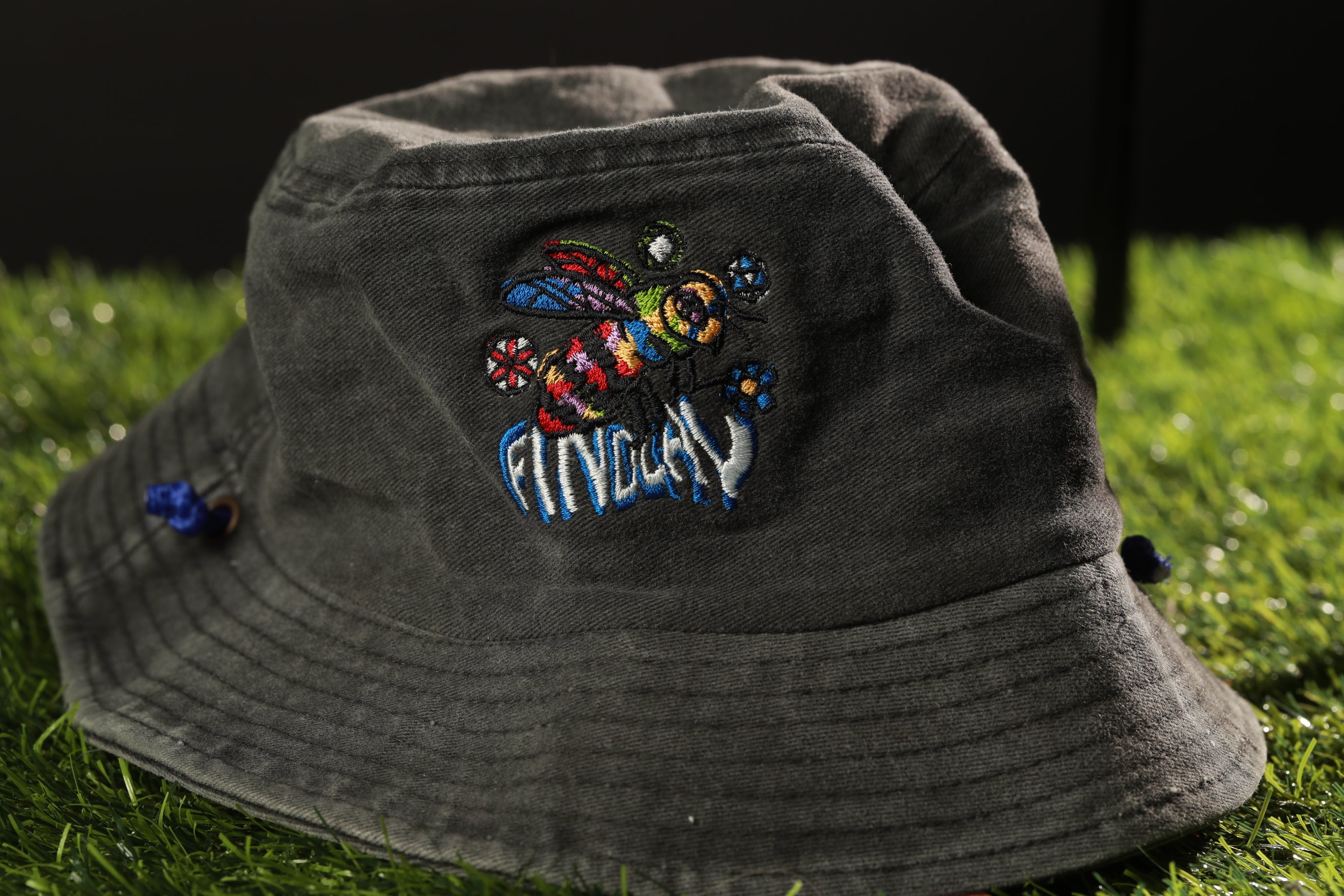 A gray bucket hat with a colorful bee embroidered on it, placed on a grassy surface.