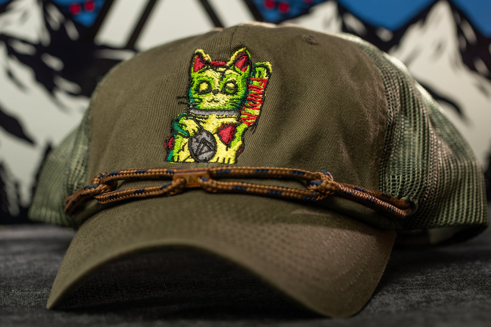 A green baseball cap with a colorful cat embroidered on it, featuring a cat-like design with a raised paw and a green and red color scheme.