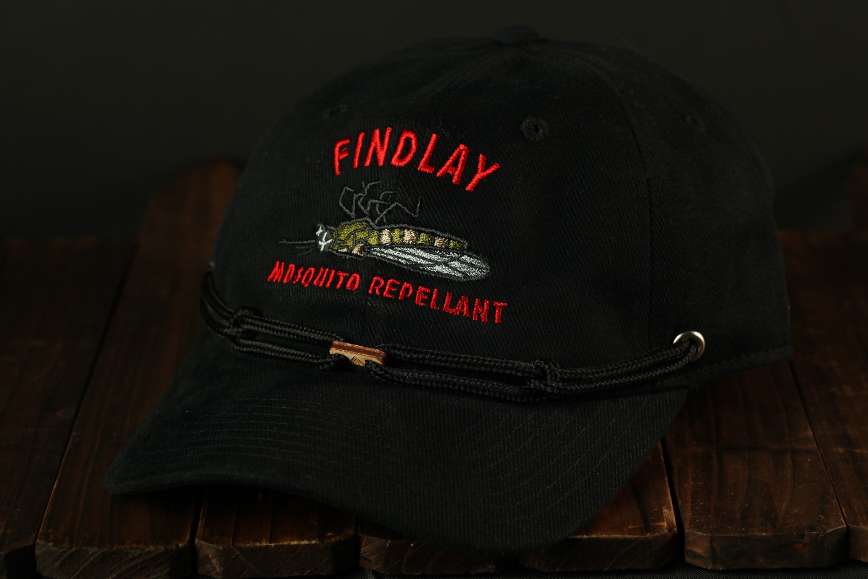 A black baseball cap with a red and white embroidered design on the front, featuring a large insect and the text "FINDLAY" and "MUSQUITO REPELLANT".