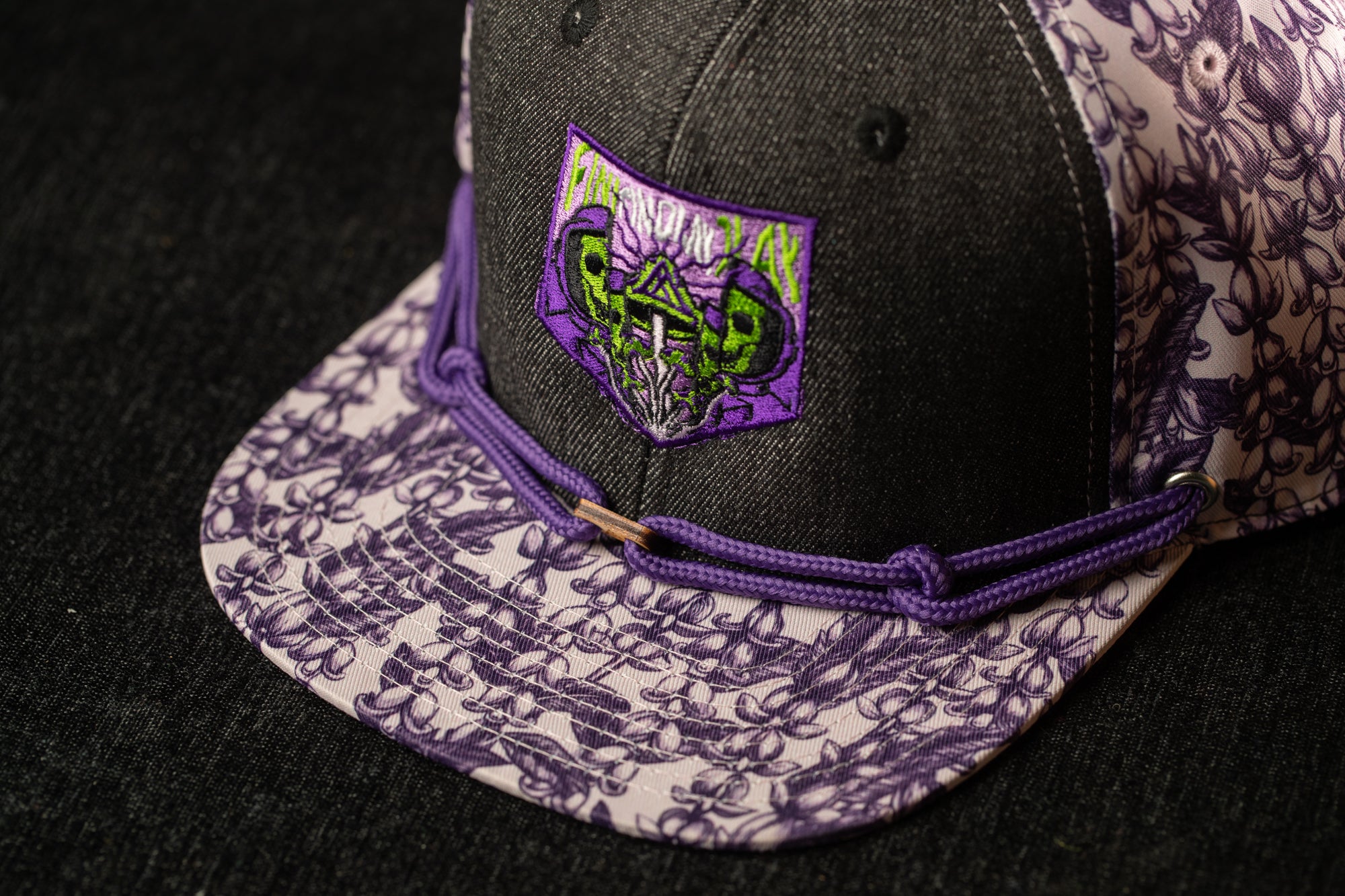 A close-up view of a baseball cap with a purple and white floral pattern on the brim and a purple and green graphic patch on the front.