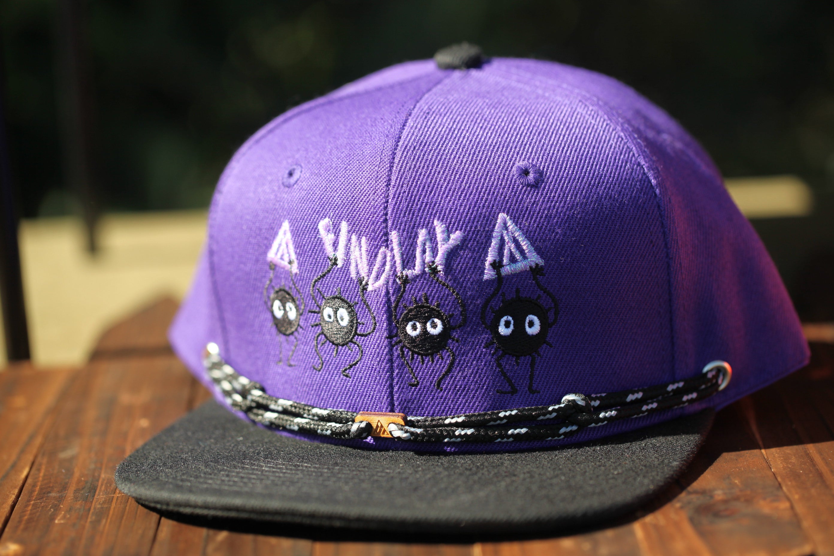 A purple baseball cap with a black brim and a black strap, featuring a design of cartoon-like black creatures with eyes and legs.