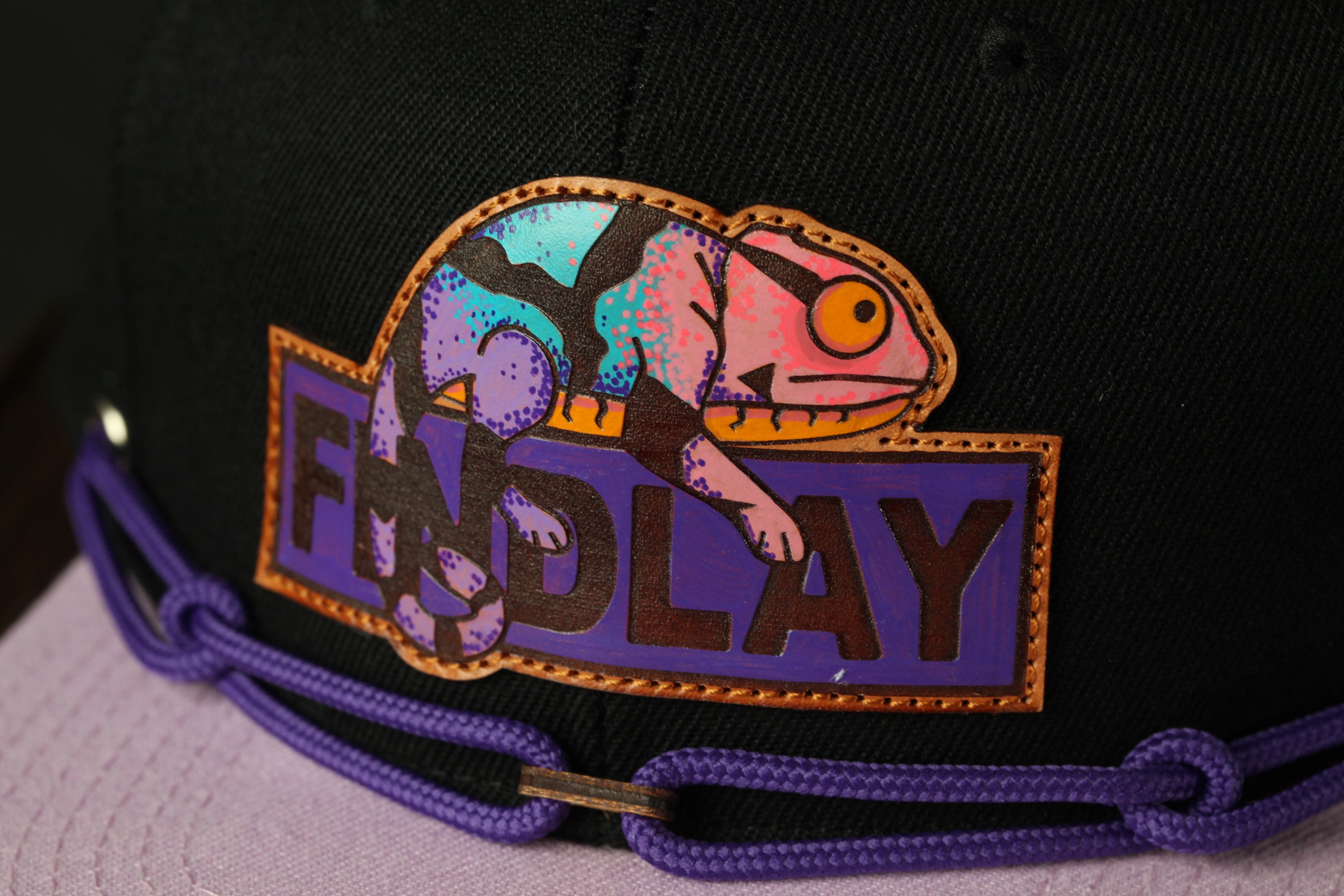 A close-up of a black baseball cap with a colorful, hand-painted chameleon design on the front.