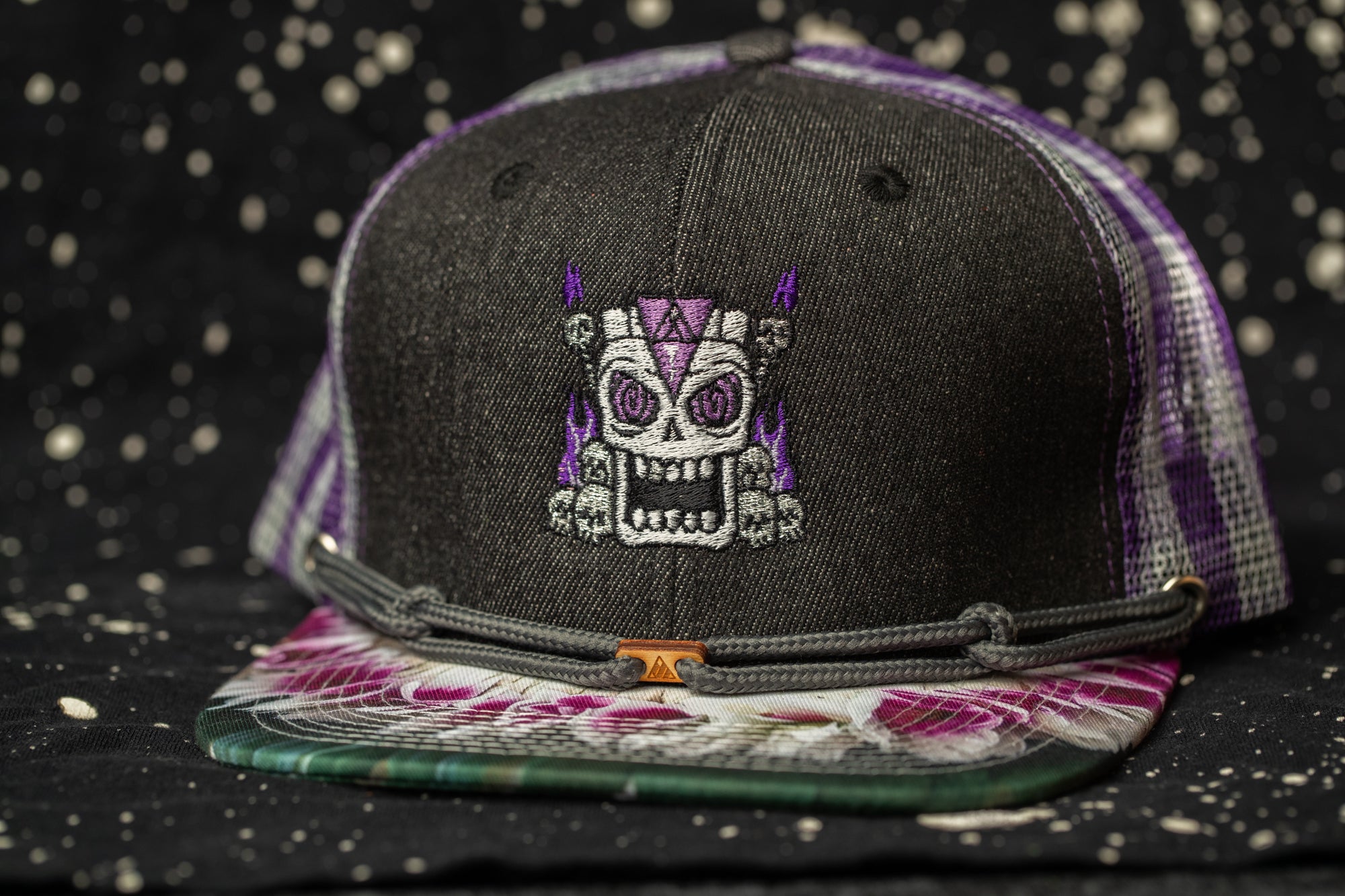 A black baseball cap with a purple skull embroidered on it, placed on a dark background with white speckles.