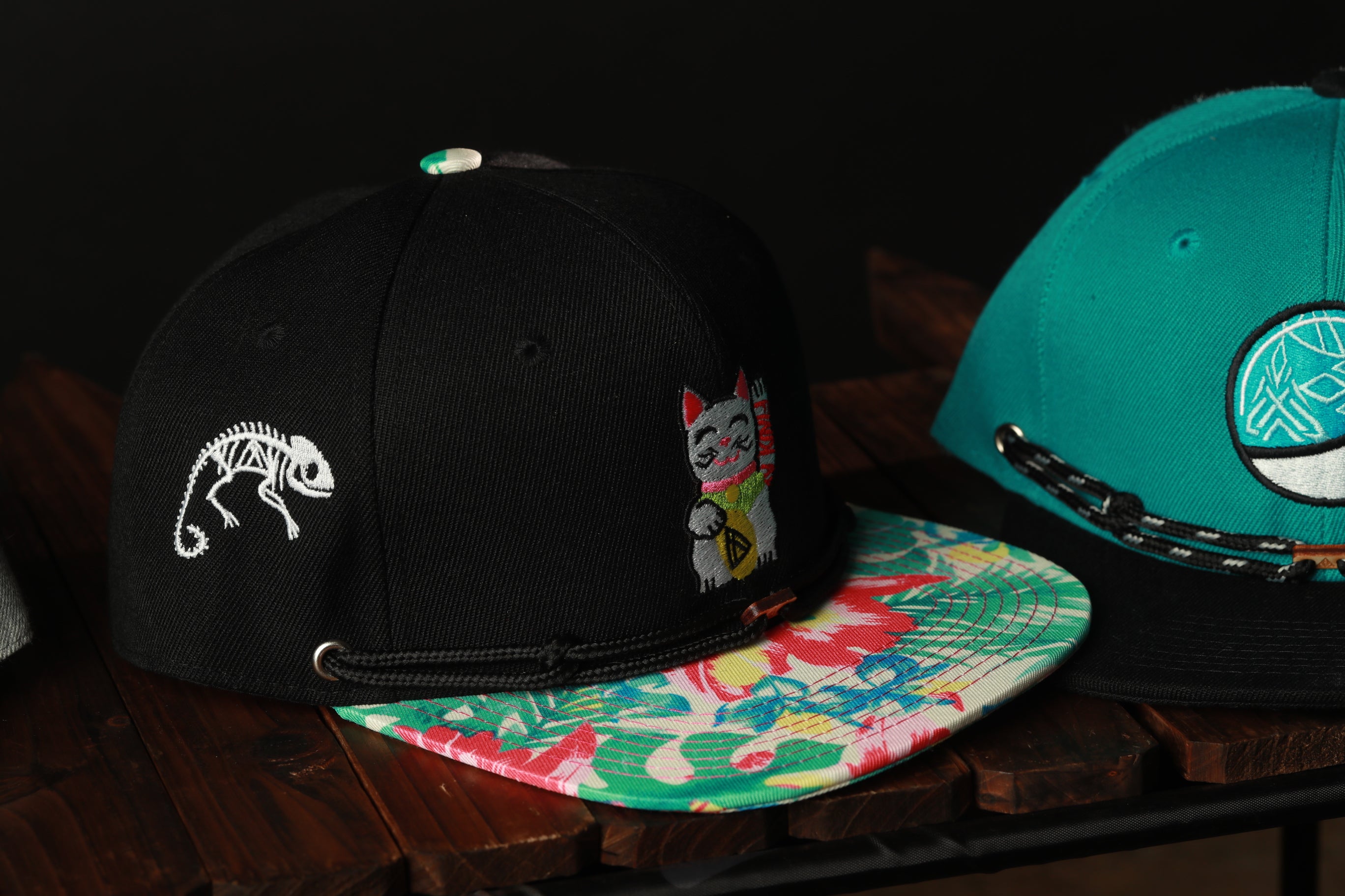 two baseball caps on a wooden surface, one black and one teal, with floral patterns on the caps.