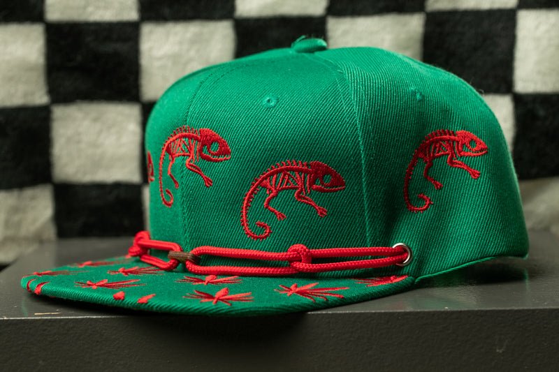 A green baseball cap with red chameleons printed on it, sitting on a gray surface against a black and white checkered background.