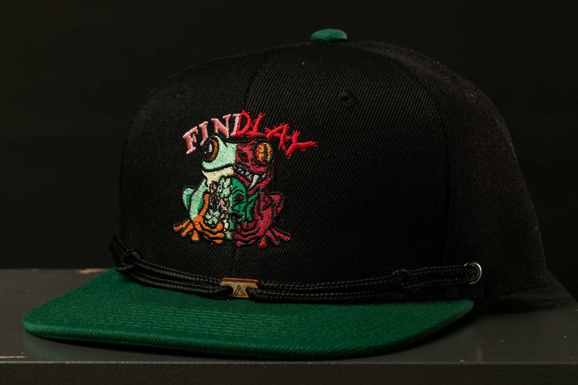 A black baseball cap with a green brim and a colorful frog embroidered on the front.