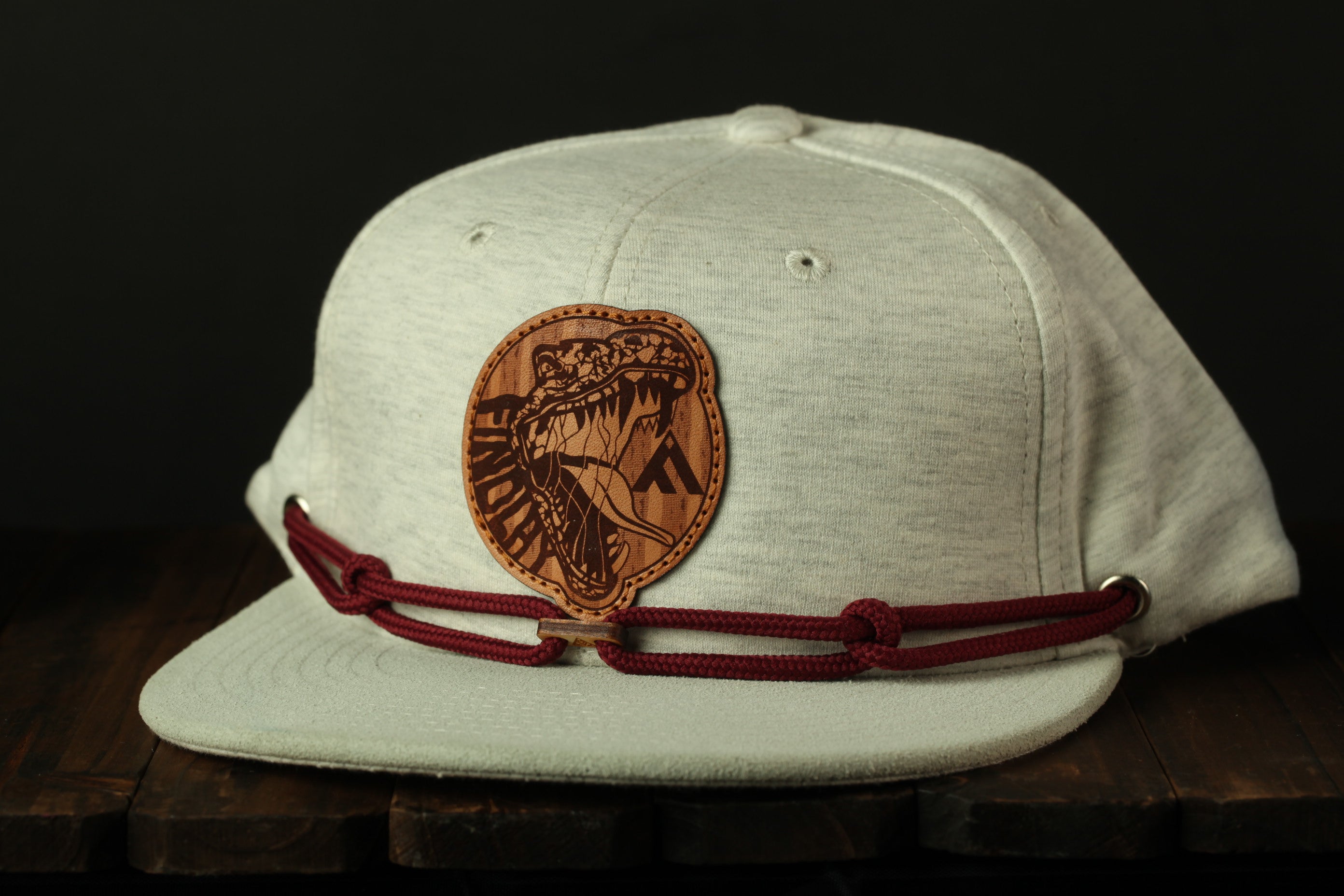 A white baseball cap with a red braided band and a circular patch featuring a stylized dinosaur or prehistoric creature on the front.