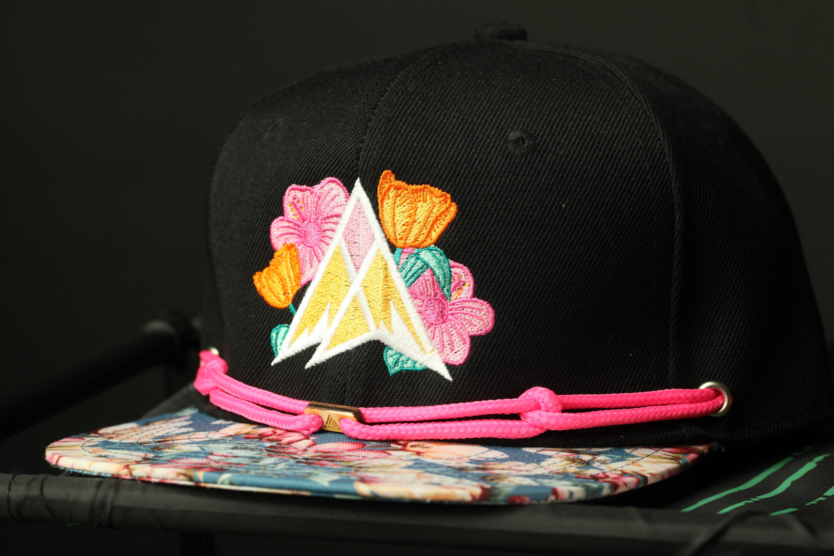 A black baseball cap with a floral embroidered logo on the front, featuring a yellow triangle and pink flowers.