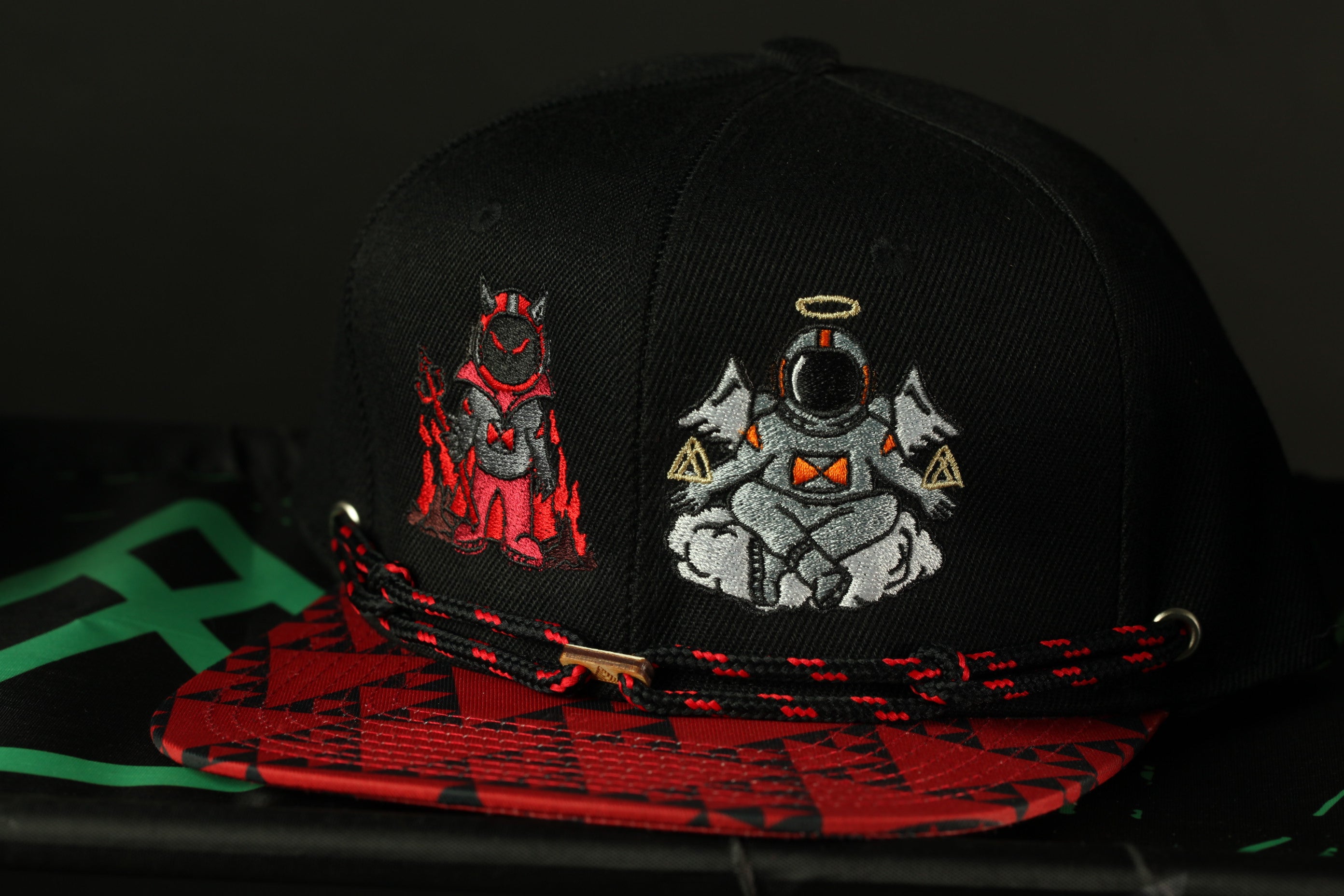 A black baseball cap with two distinct embroidered characters on it, one in red and one in white, sitting on a red and black patterned fabric.