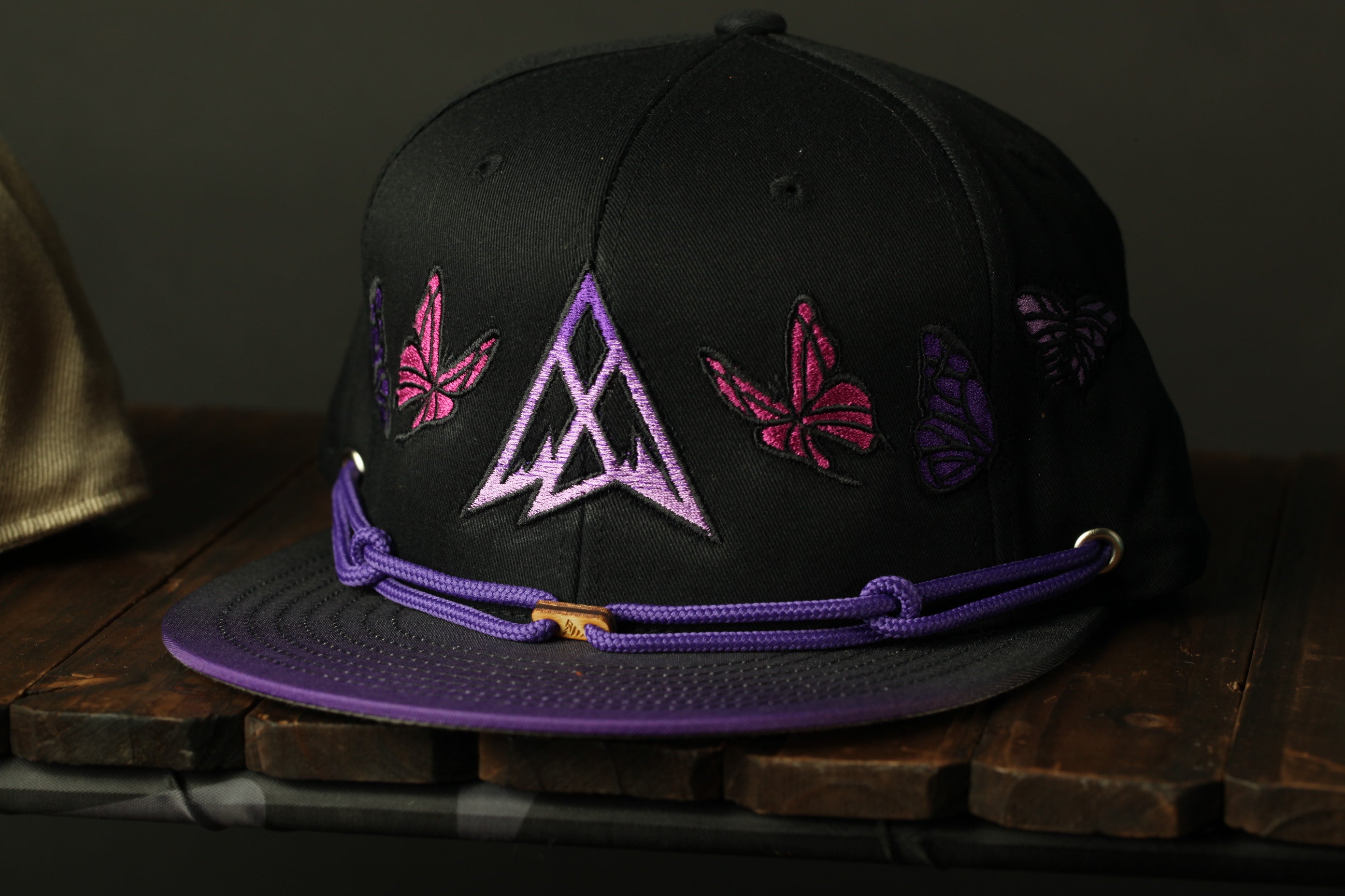 A black baseball cap with purple and pink butterfly embroidery, sitting on a wooden surface.