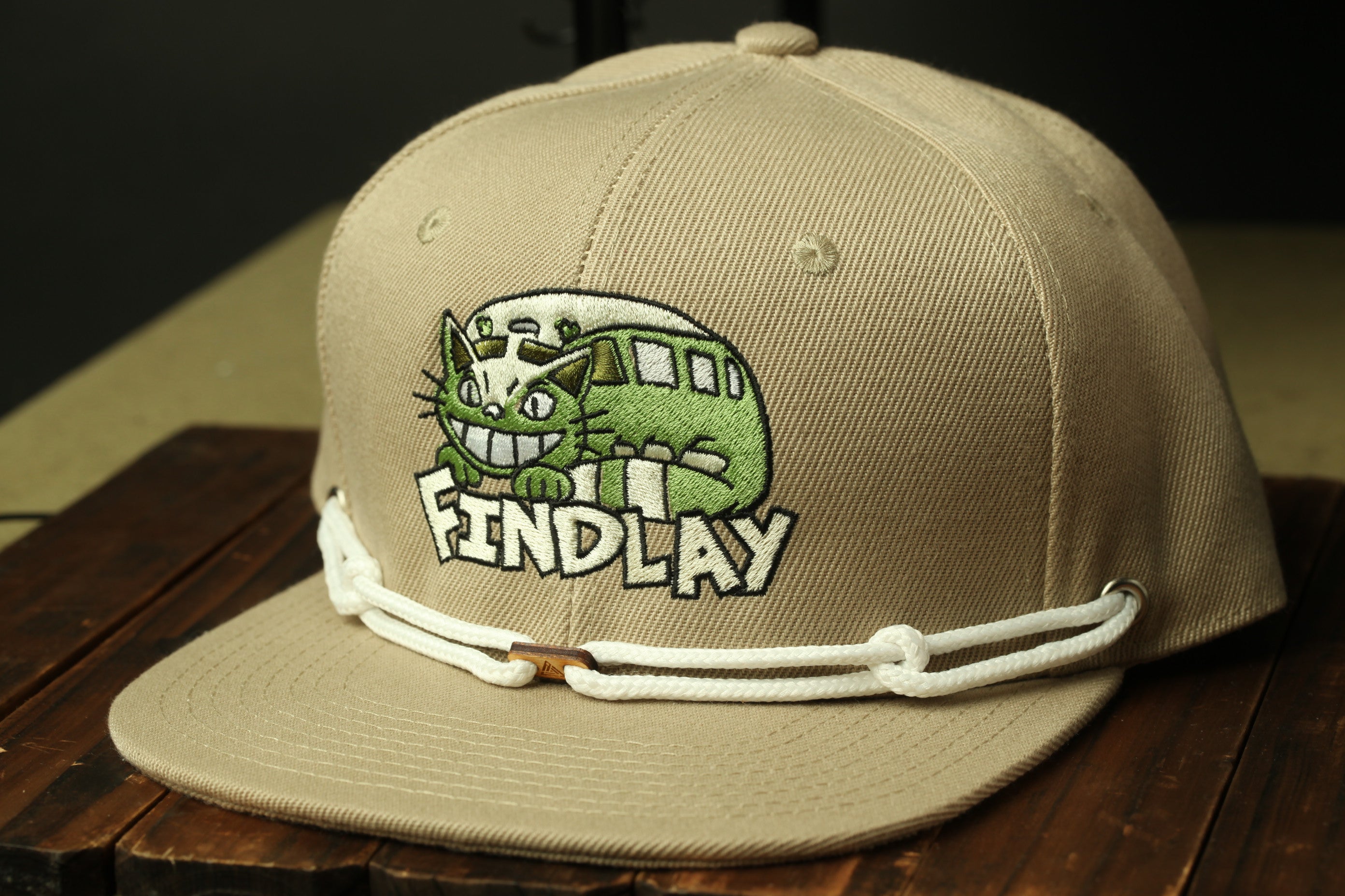 A beige baseball cap with a cartoon cat design and the word "FINDLAY" printed on it.