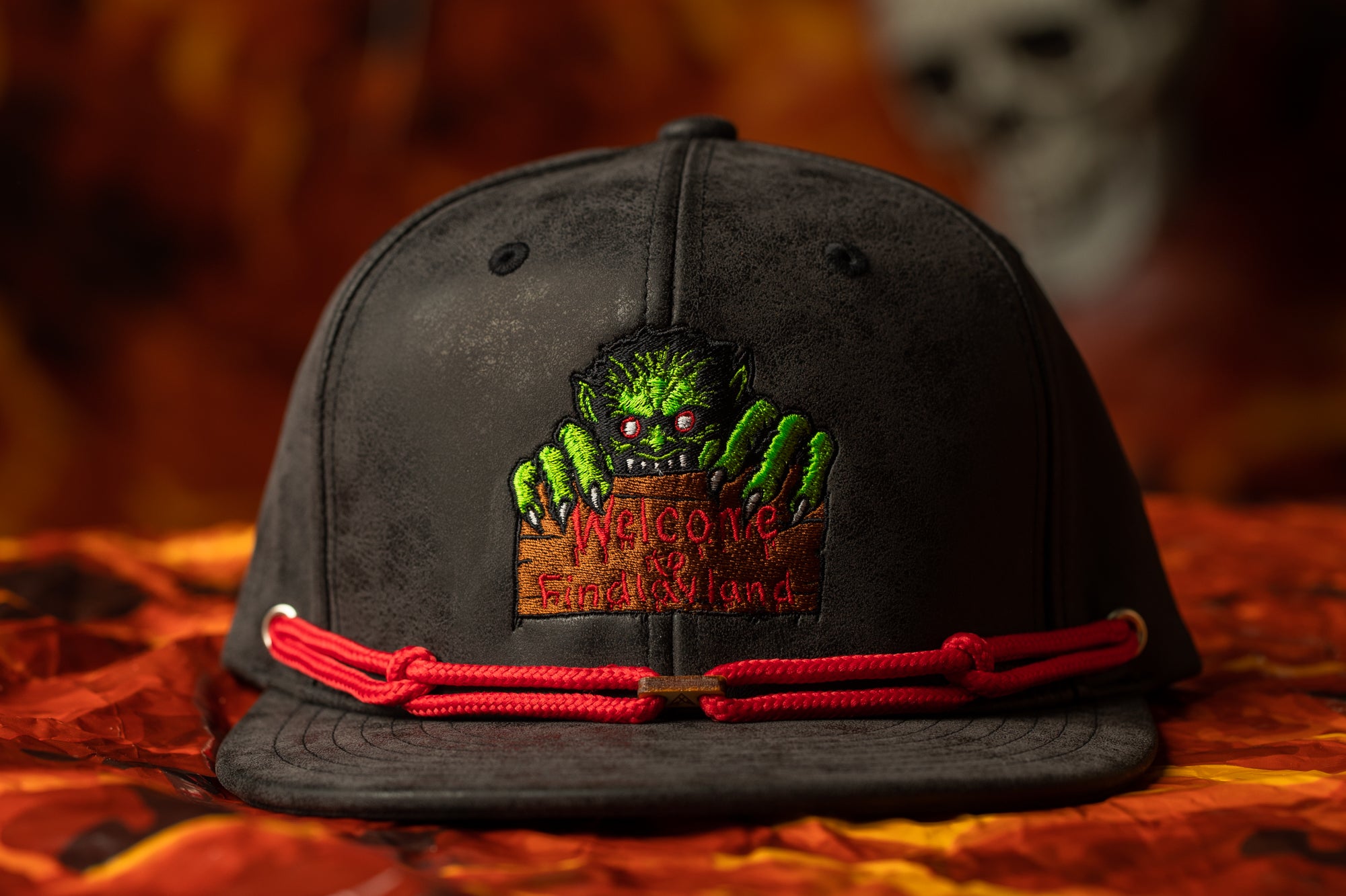 A black baseball cap with a red string and a green monster graphic on it, placed on a red and orange patterned surface.