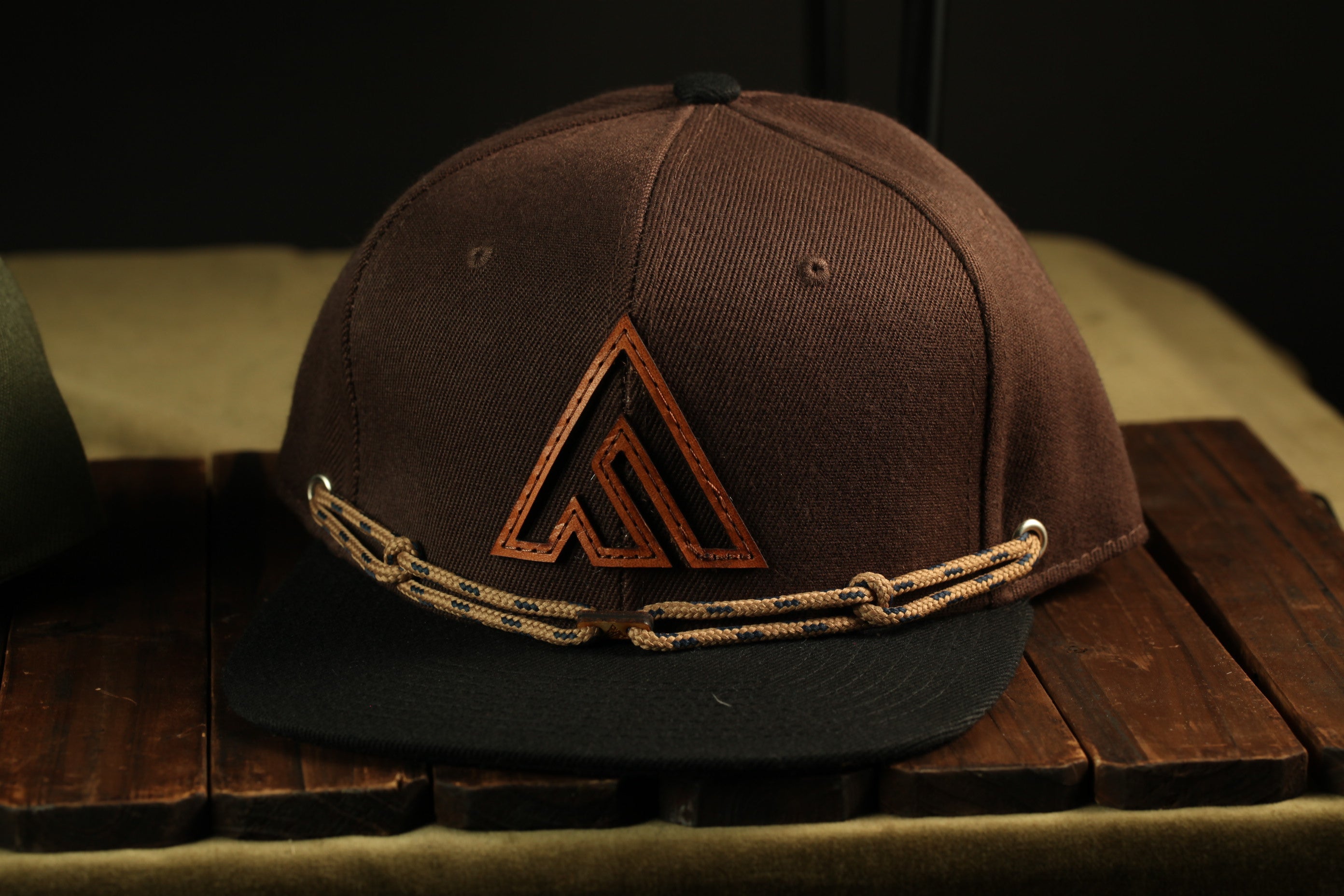 A brown baseball cap with a black brim and a brown rope-like strap is displayed on a wooden surface.