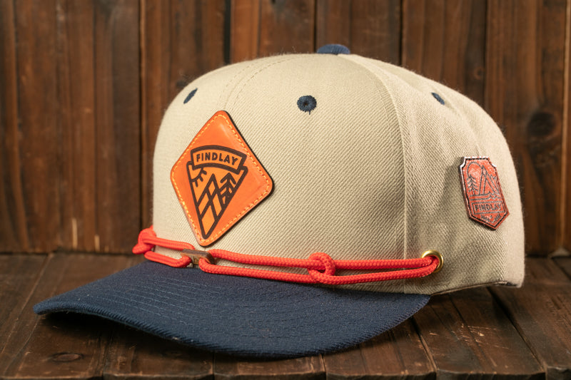 A beige baseball cap with a blue brim and orange and red accents, including a logo patch and a red string.