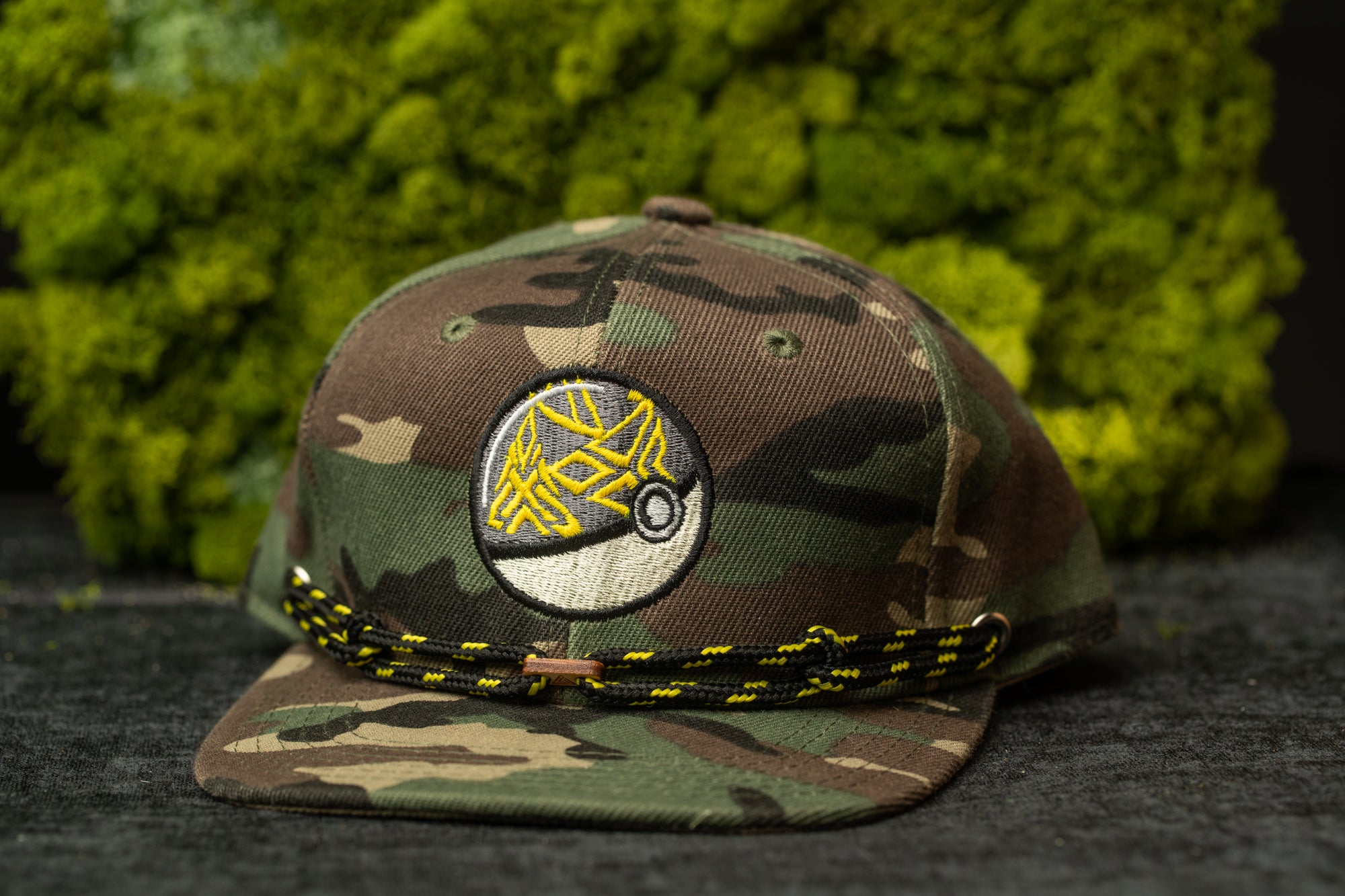 A camouflage baseball cap with a circular logo on the front, placed on a dark surface against a background of green moss.