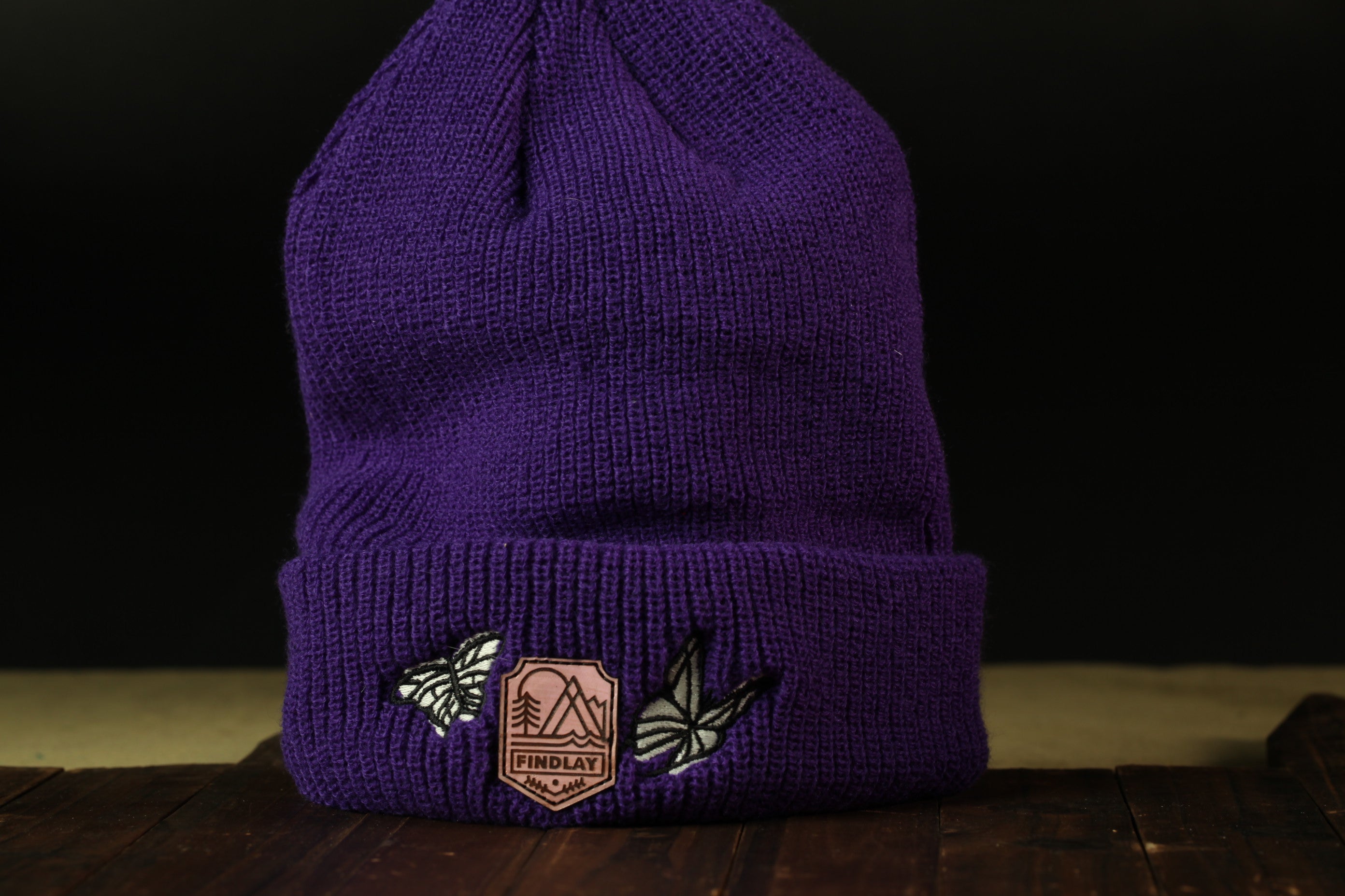 A purple knit beanie with a patch featuring a mountain and butterfly design, placed on a wooden surface against a dark background.