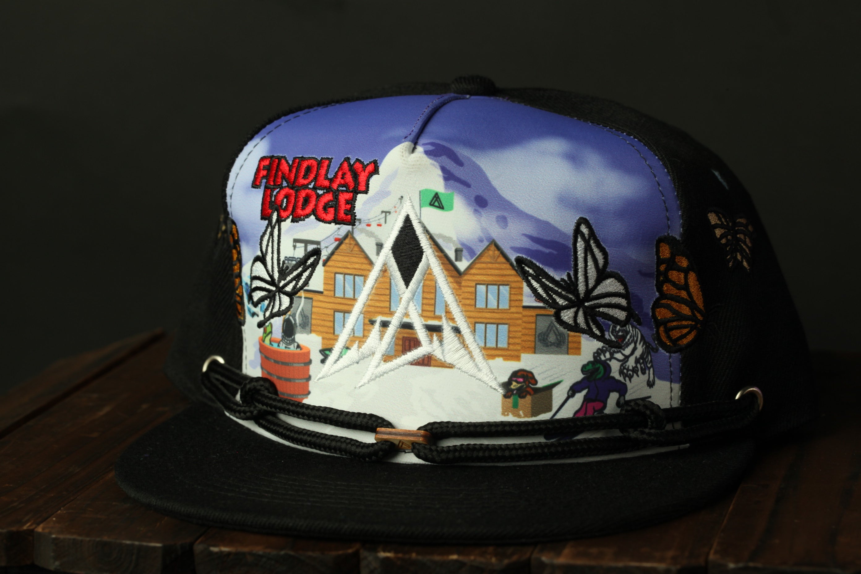 A black baseball cap with a colorful graphic design depicting a lodge, butterflies, and a mountain.