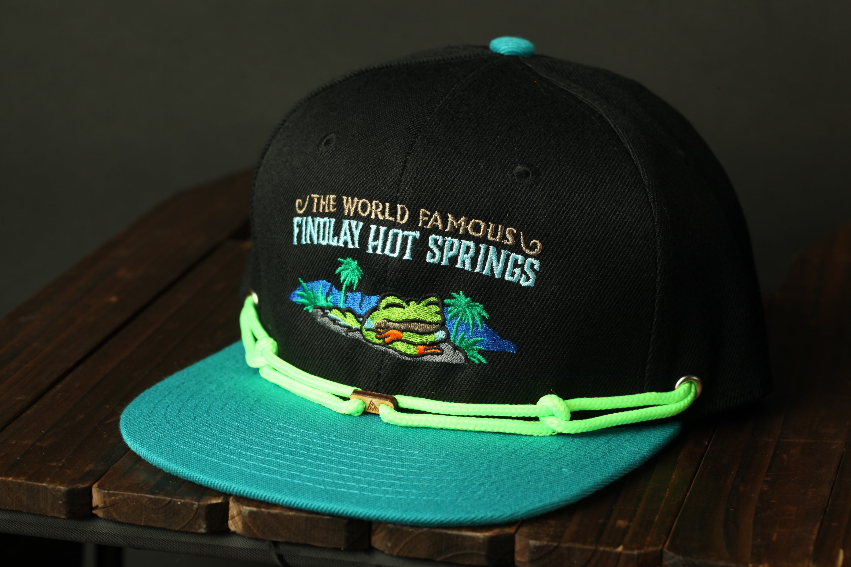A black baseball cap with a teal brim and a graphic of a tropical island on the front, sitting on a wooden surface.