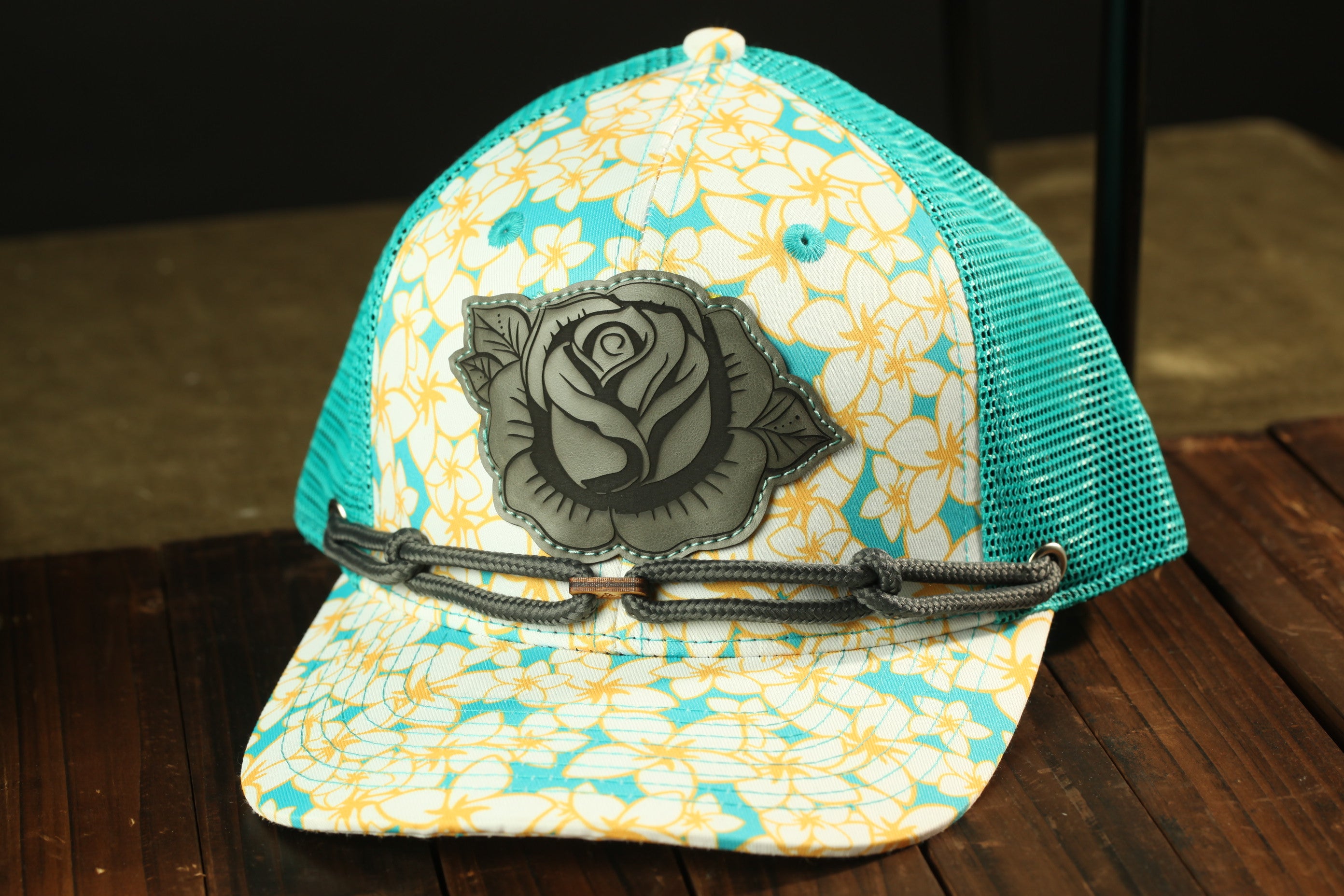 A turquoise and white floral baseball cap with a rose design on the front, sitting on a wooden surface.