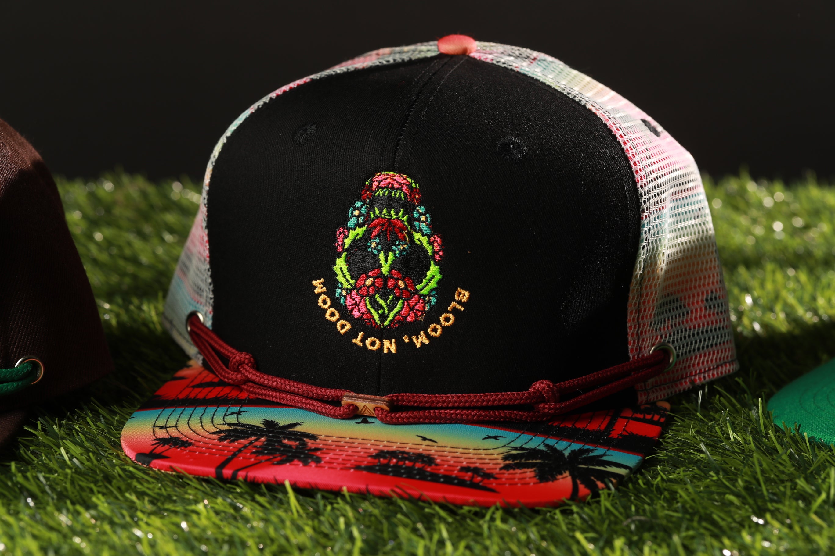 A colorful baseball cap with a floral design and the text "BLOOM" on it, placed on a grassy surface.