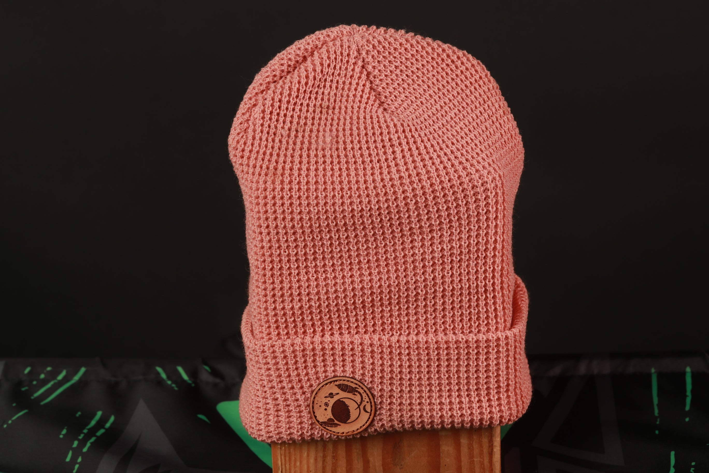 A pink knitted beanie with a circular logo on the front, placed on a wooden surface against a black background.