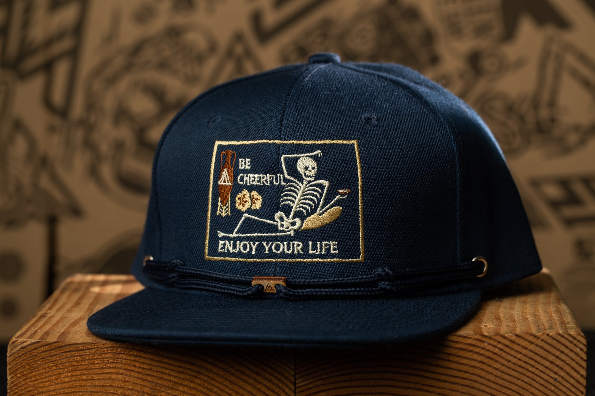 A navy blue baseball cap with a white skull and crossbones design, along with the text "Be cheerful, enjoy your life" printed on it.