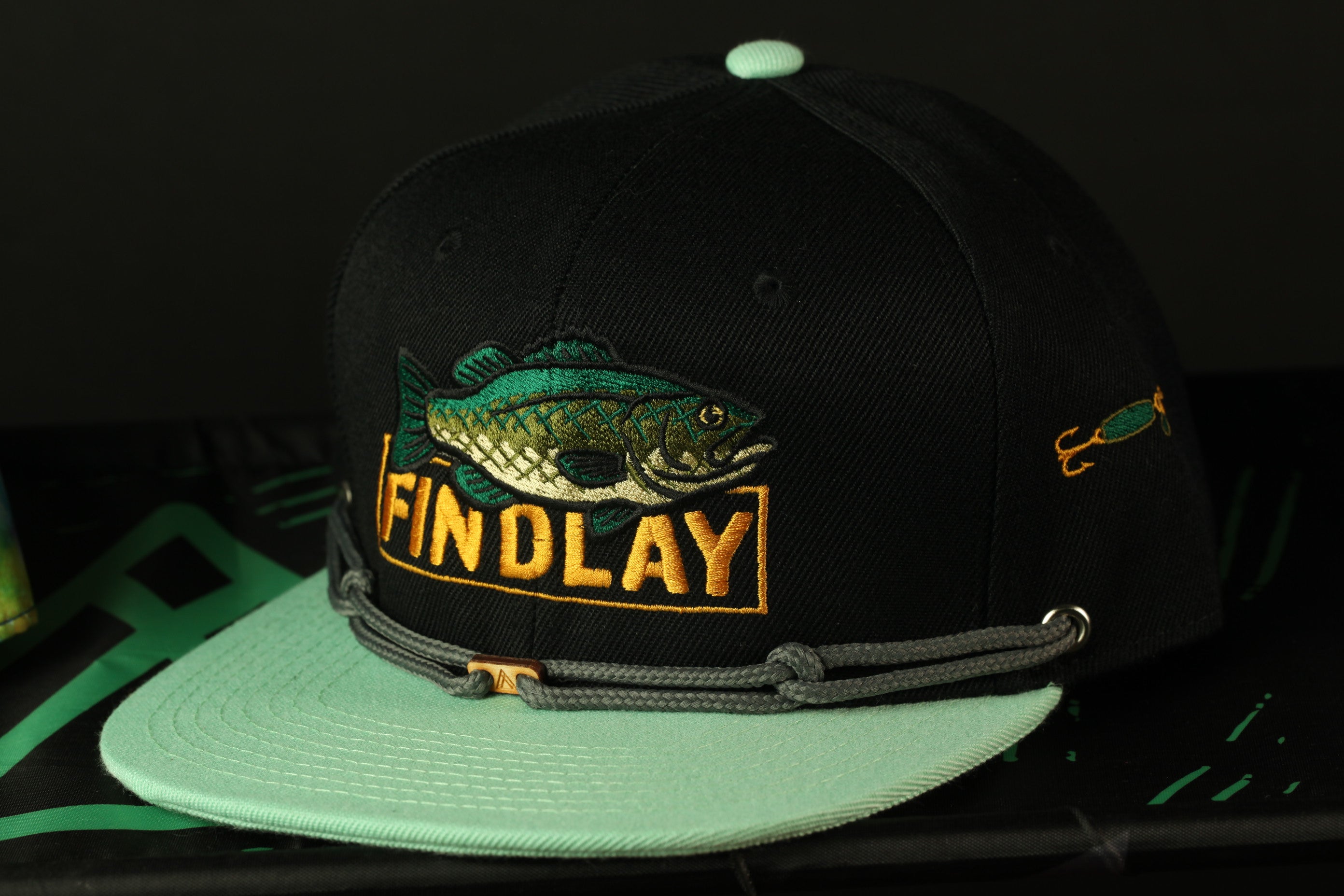 A black baseball cap with a fish embroidered on it, featuring the word "FINOLAY" in orange letters.