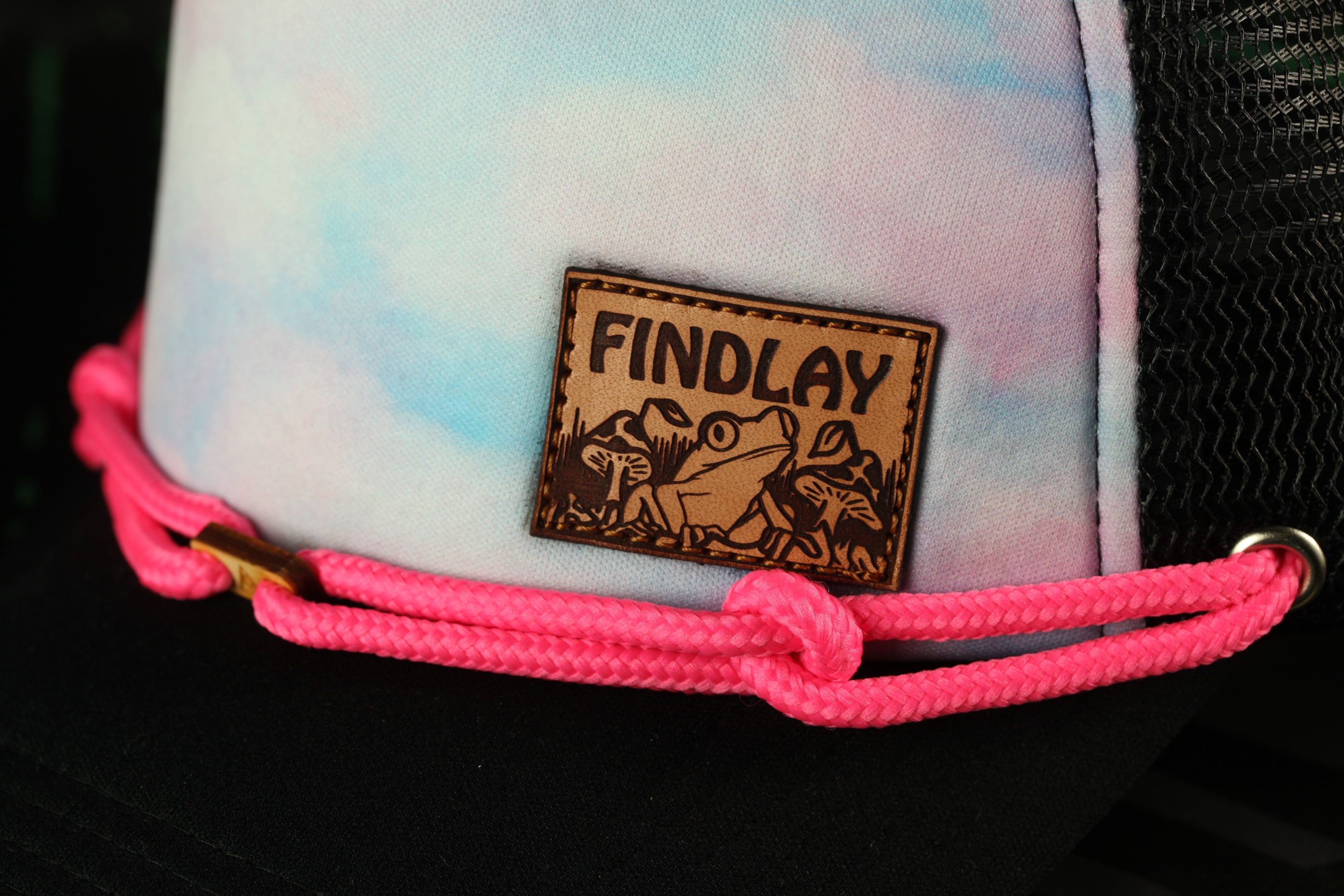 A close-up of a hat with a pink braided strap and a small leather patch on the front, featuring a frog and the word "FINDLAY".
