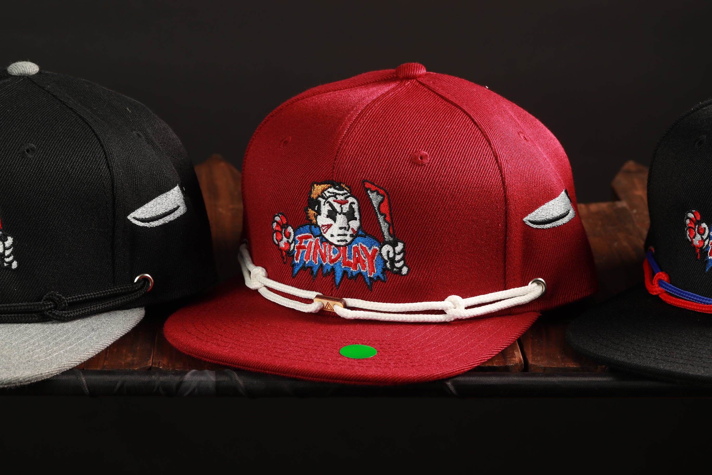 Three baseball caps are displayed on a shelf, with one red cap featuring a graphic design and the other two black caps.