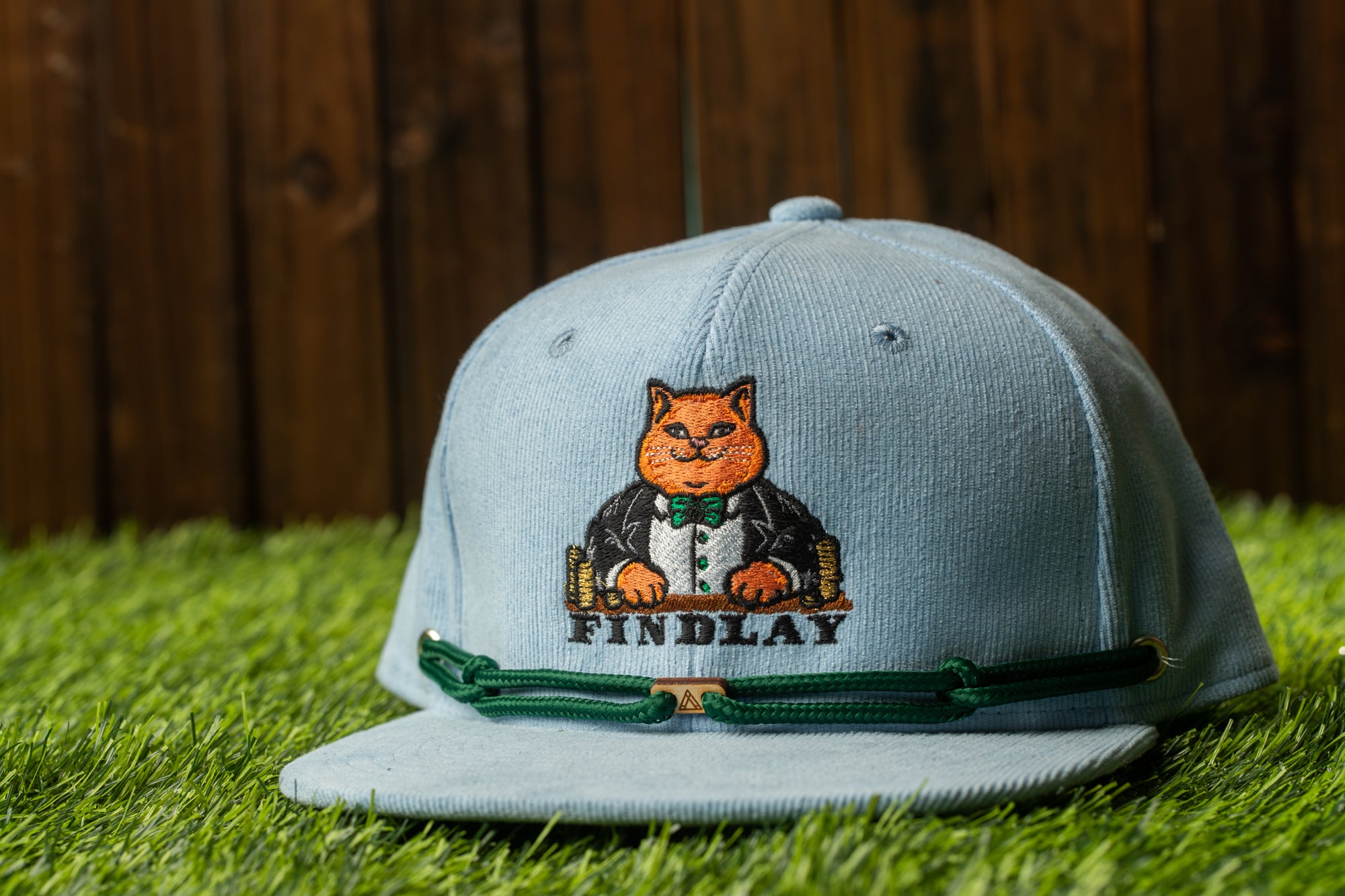 A light blue baseball cap with a cartoon cat and the text "FINDA" printed on it, placed on a grassy surface.