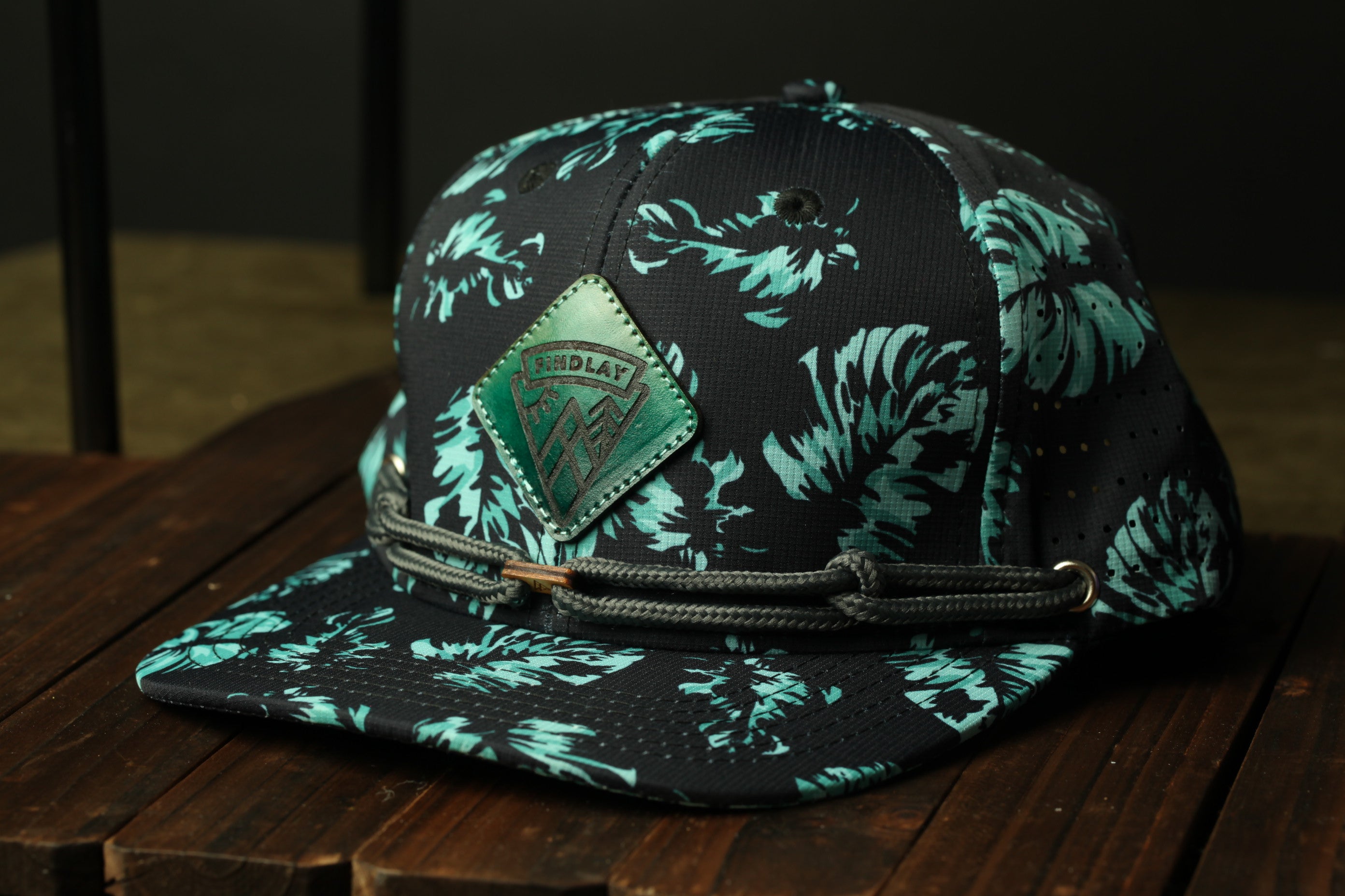 A black and teal floral baseball cap with a green patch on the front, resting on a wooden surface.