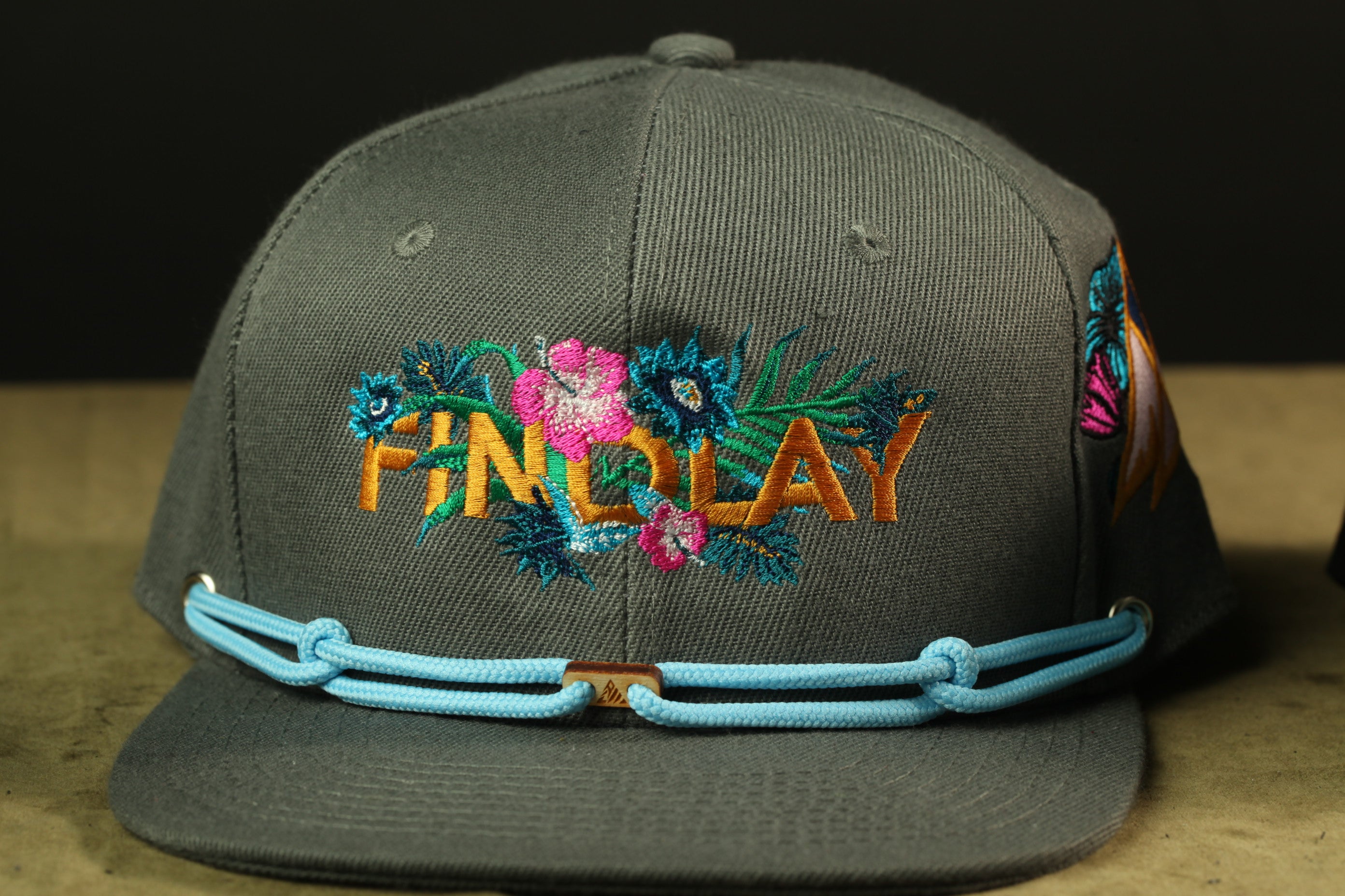 A gray baseball cap with a floral embroidered design and a blue adjustable strap.