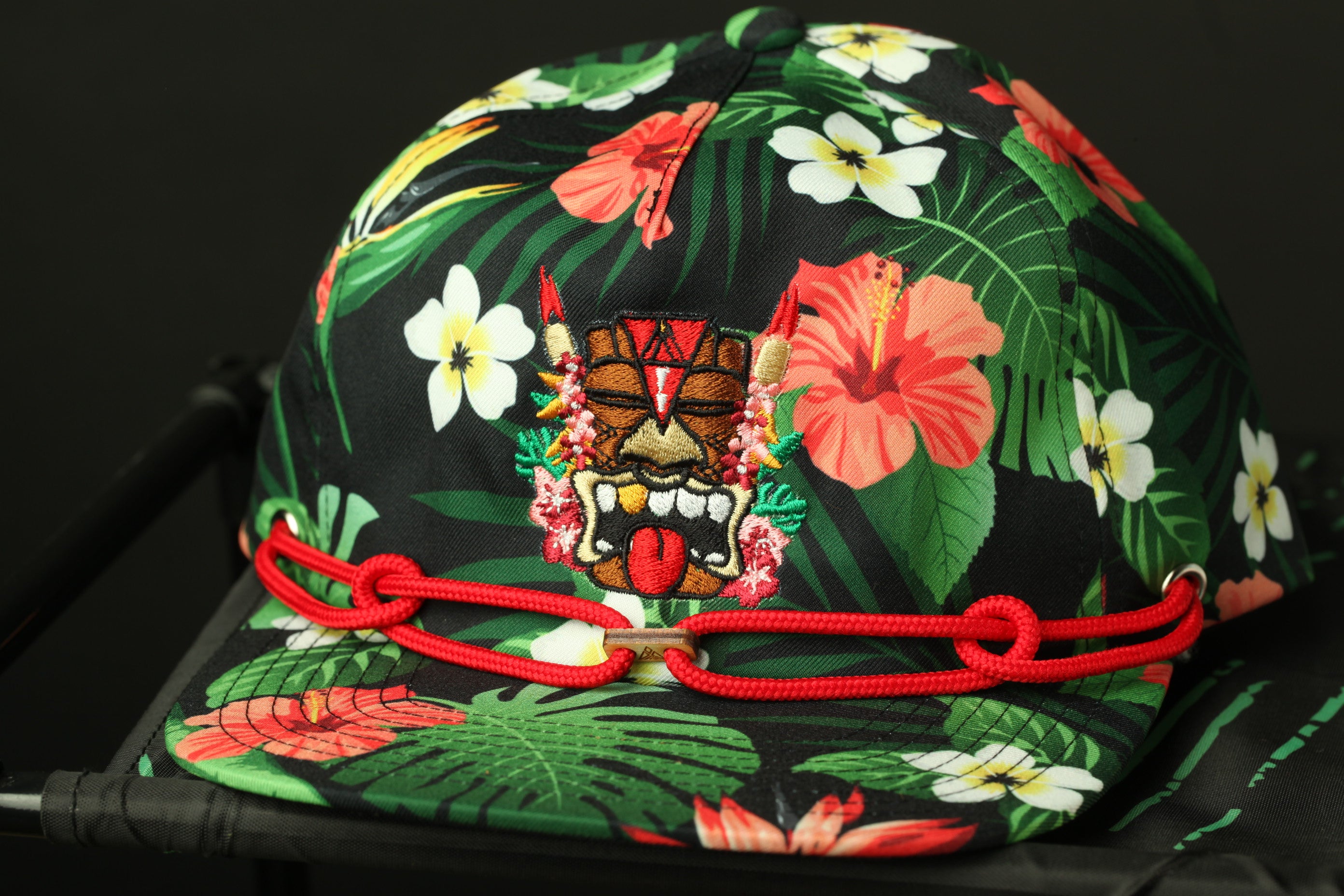 A colorful tropical-themed baseball cap with a tiki mask design and red braided straps.