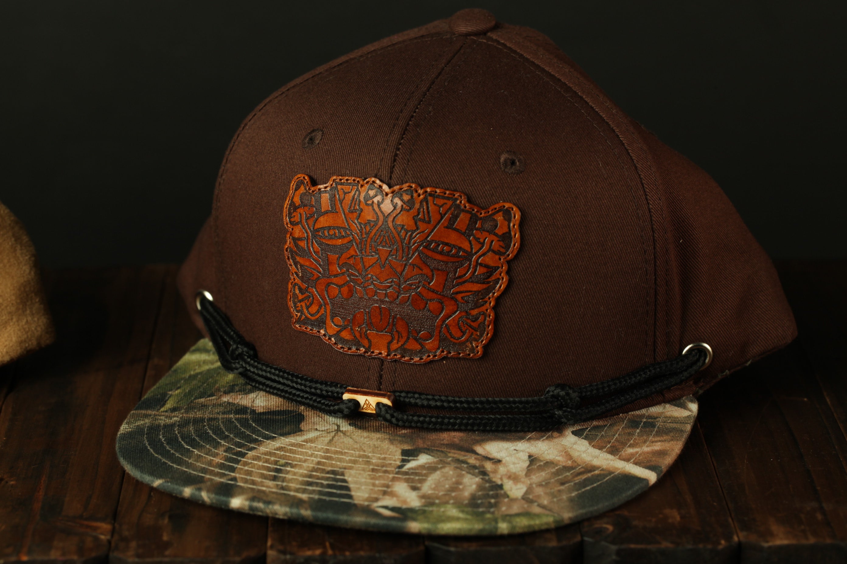 A brown baseball cap with a camouflage pattern and a leather patch on the front, placed on a wooden surface.