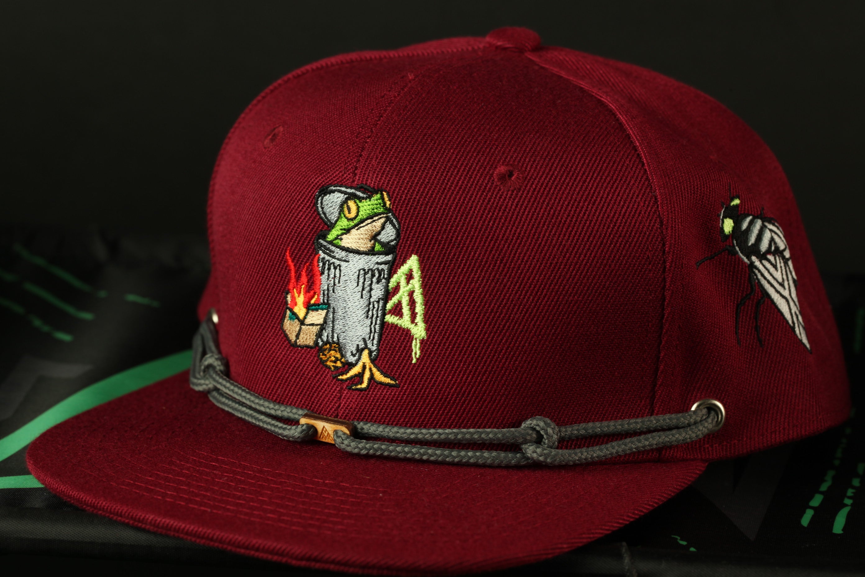 A maroon baseball cap with a unique design featuring a frog, a trash can, and a fly.