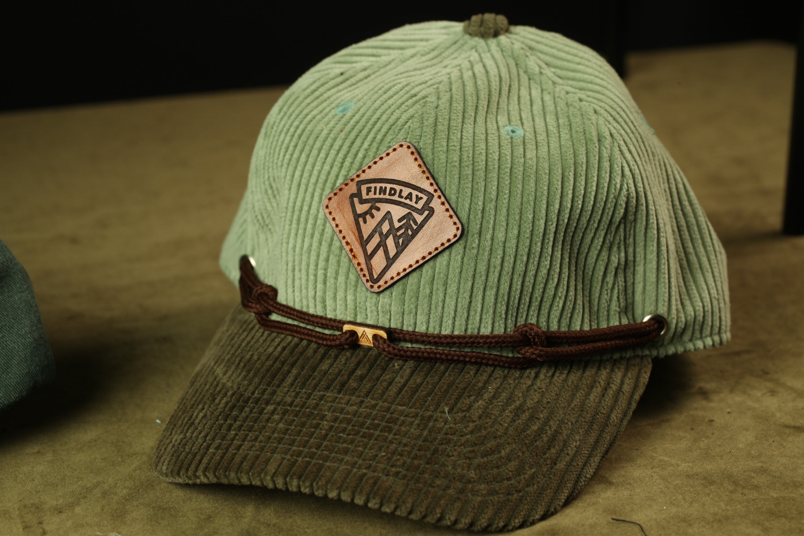 A green baseball cap with a brown leather patch and a brown leather strap, placed on a brown surface.