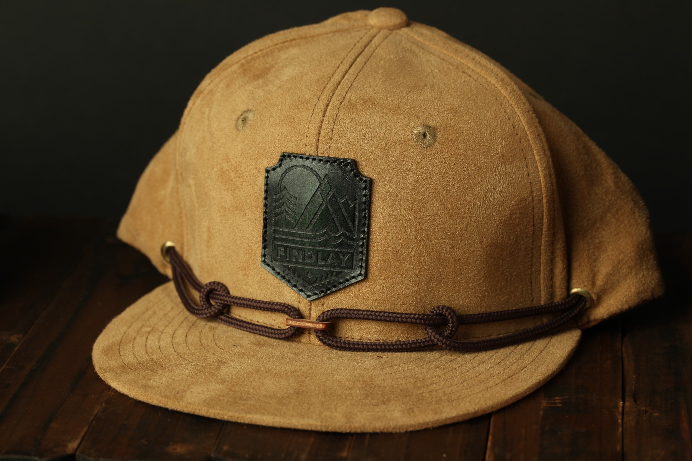 A tan baseball cap with a black leather patch and brown leather straps, placed on a wooden surface.