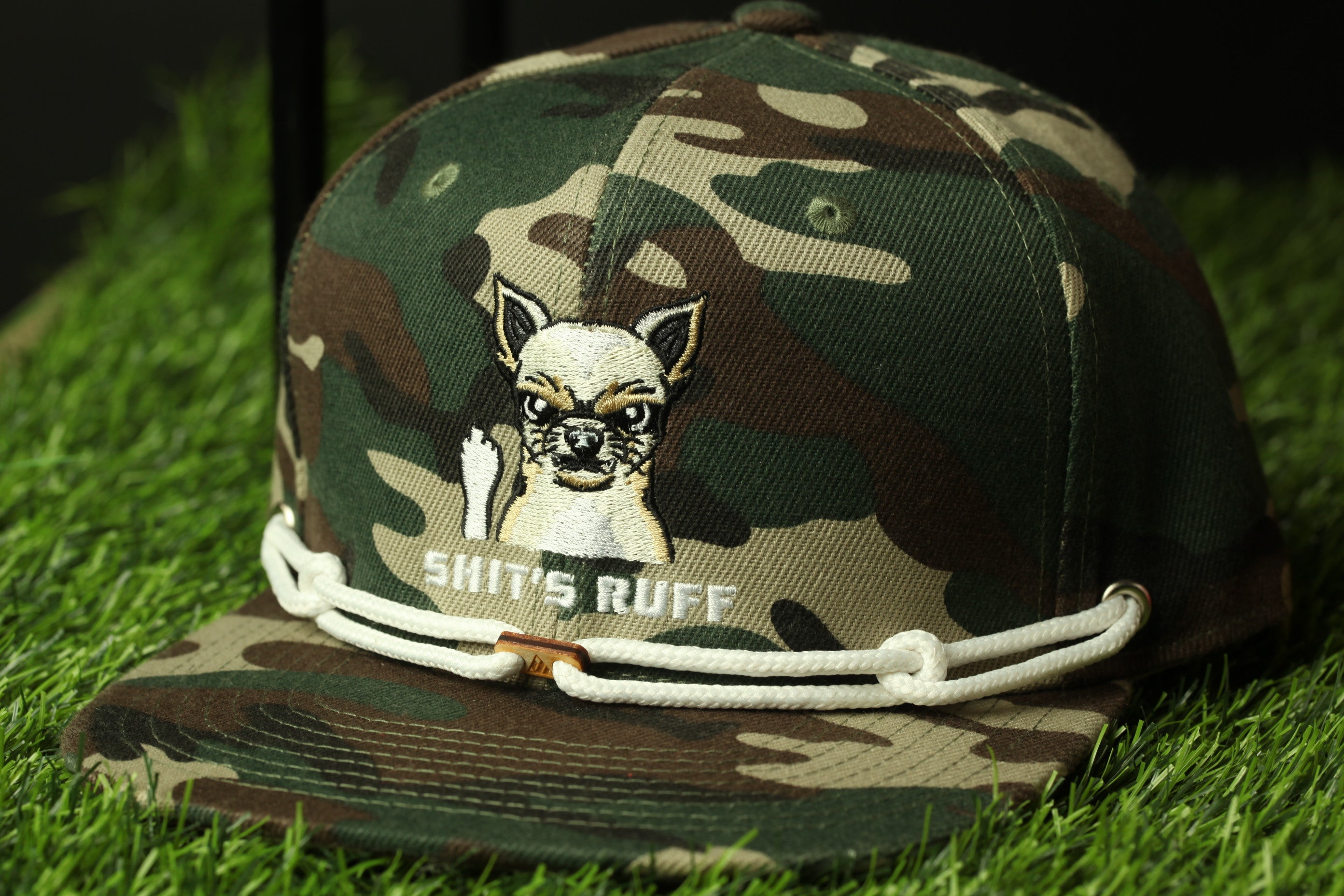 A camouflage baseball cap with a cartoon dog and the text "SHIT'S RUFF" printed on it, sitting on a grassy surface.