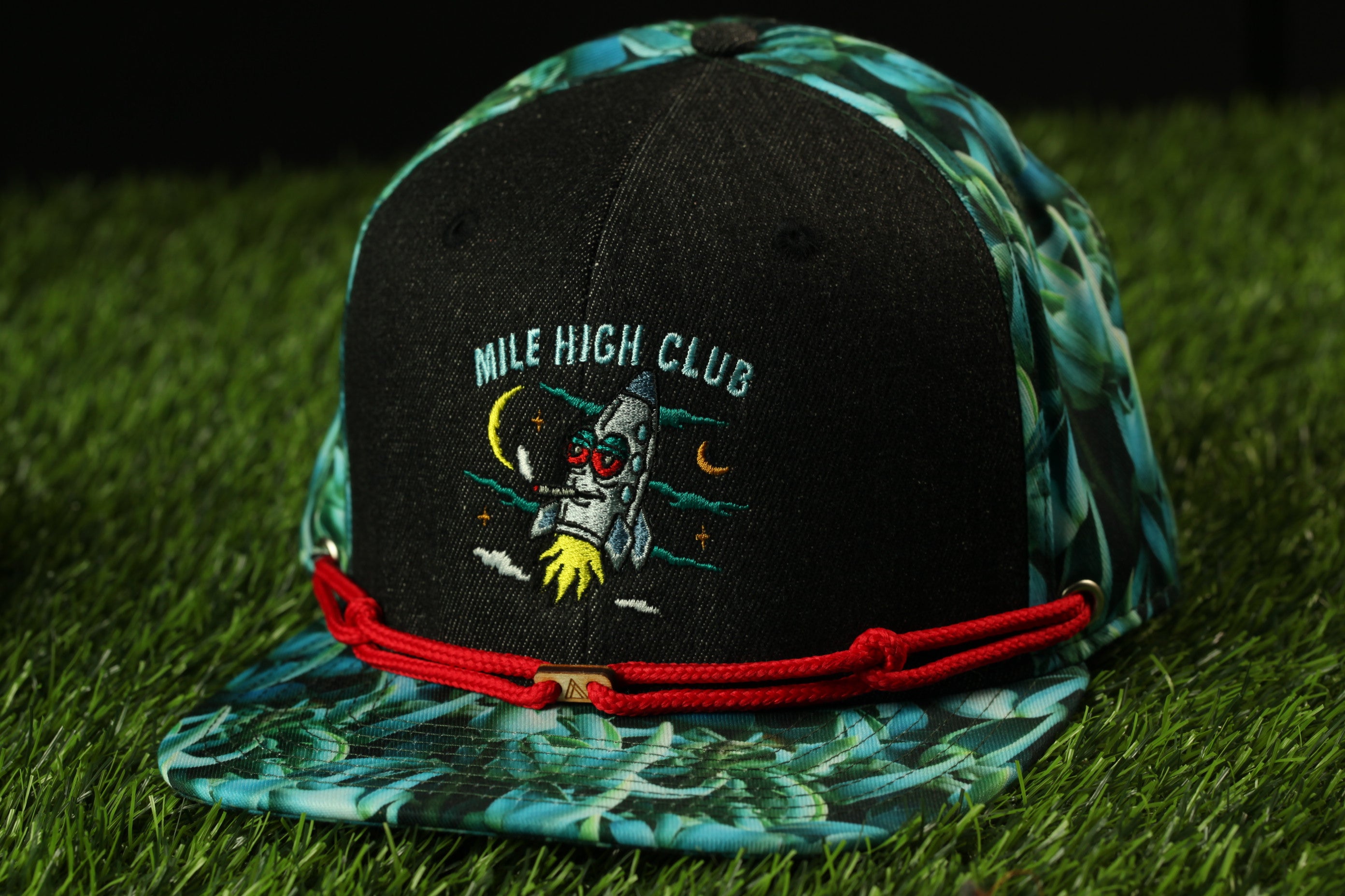 A black baseball cap with a tropical print pattern and a red string around the brim, featuring an embroidered rocket ship design.