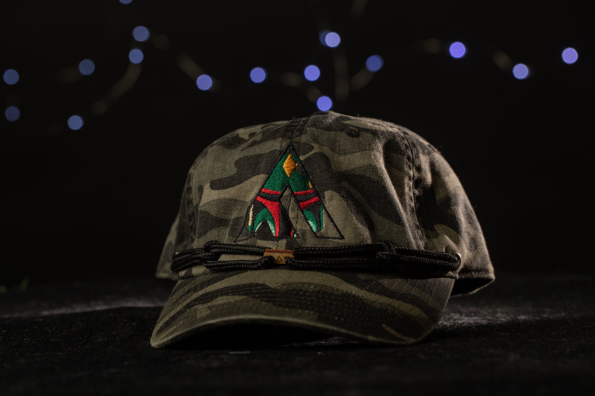 A camouflage baseball cap with a colorful embroidered logo on the front, set against a dark background with twinkling lights.