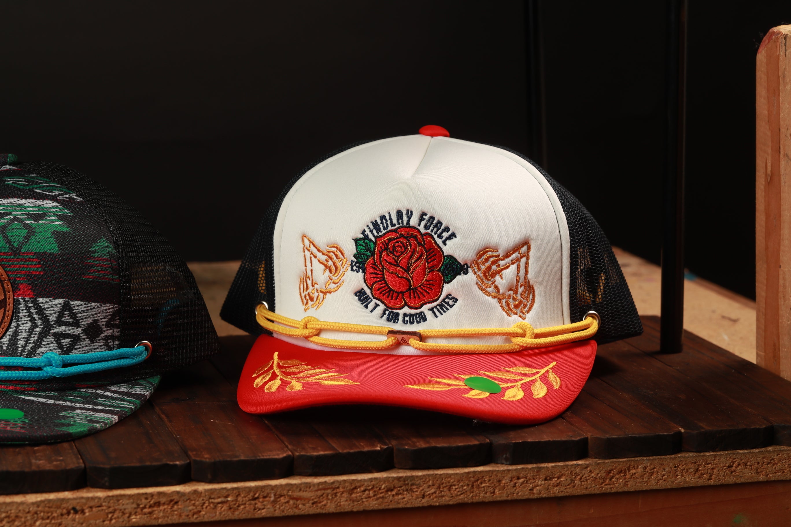 A white baseball cap with a red and gold design, featuring a red rose and gold wings, is displayed on a wooden surface.